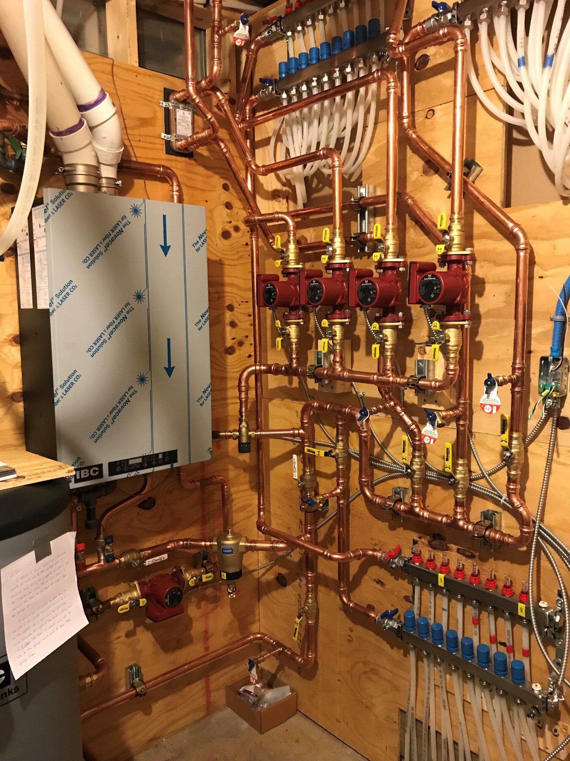 Copper pipes and valves of a hydronic heating system, installed on a wooden wall. A water heater is on the left.