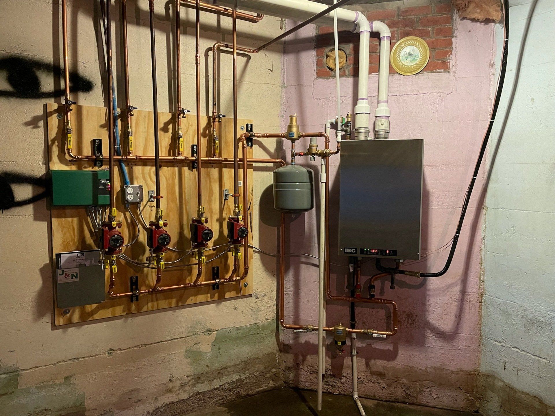 Heating system with copper pipes, pumps, and a boiler in a basement corner. A wooden board and brick wall are visible.