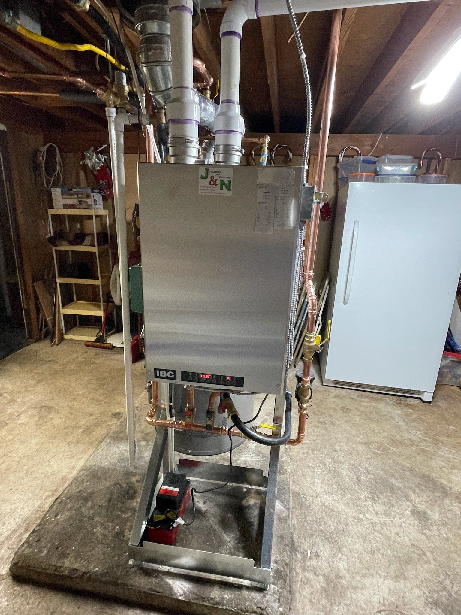 A silver boiler with copper pipes in a utility room. White PVC pipes are overhead. The floor is concrete.