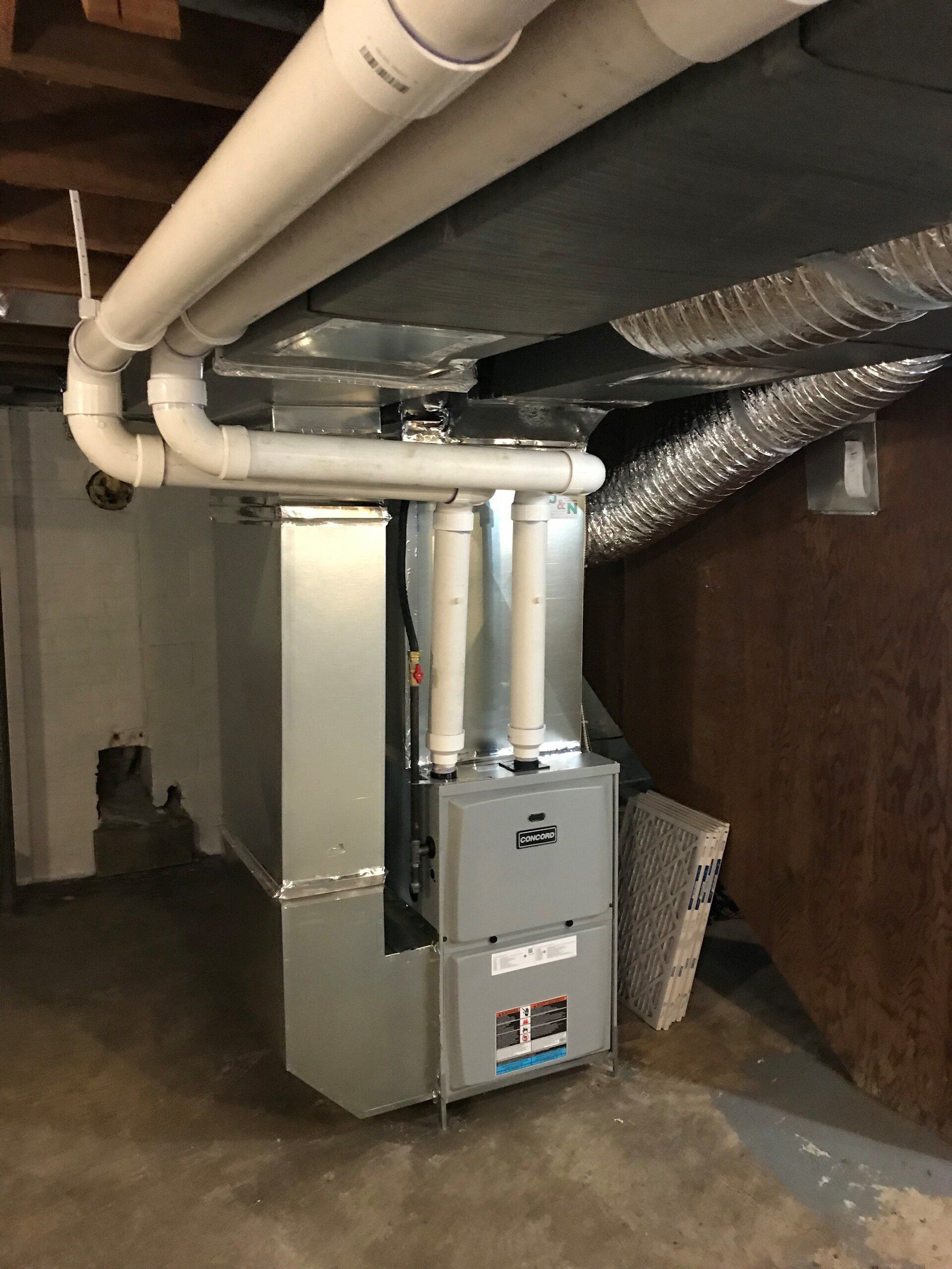 Furnace in a basement. White PVC pipes connect to the unit and run along the ceiling. A filter is visible on the side.