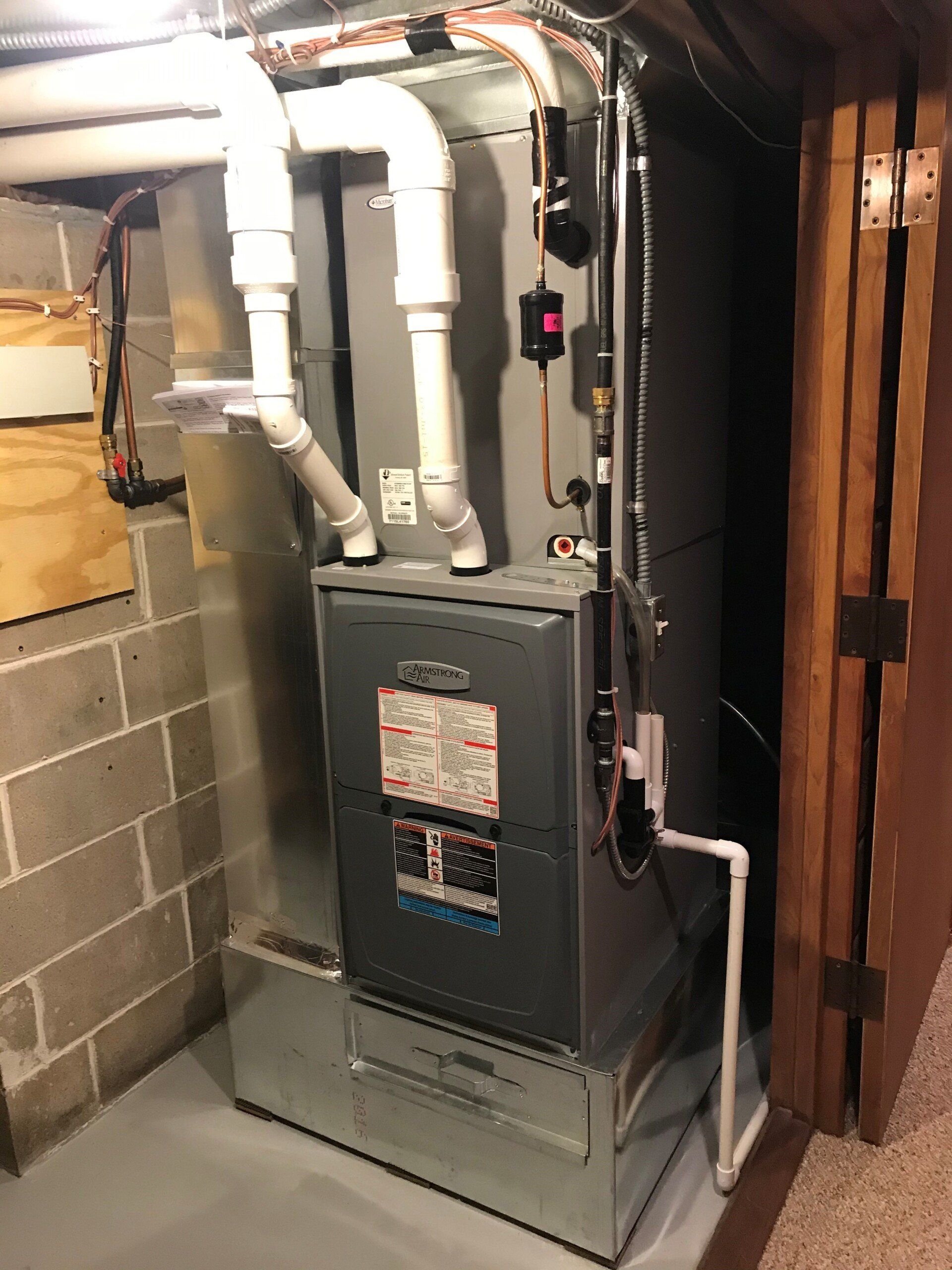 A gas furnace is installed in a basement. It has white PVC pipes attached and is located next to a wooden door.