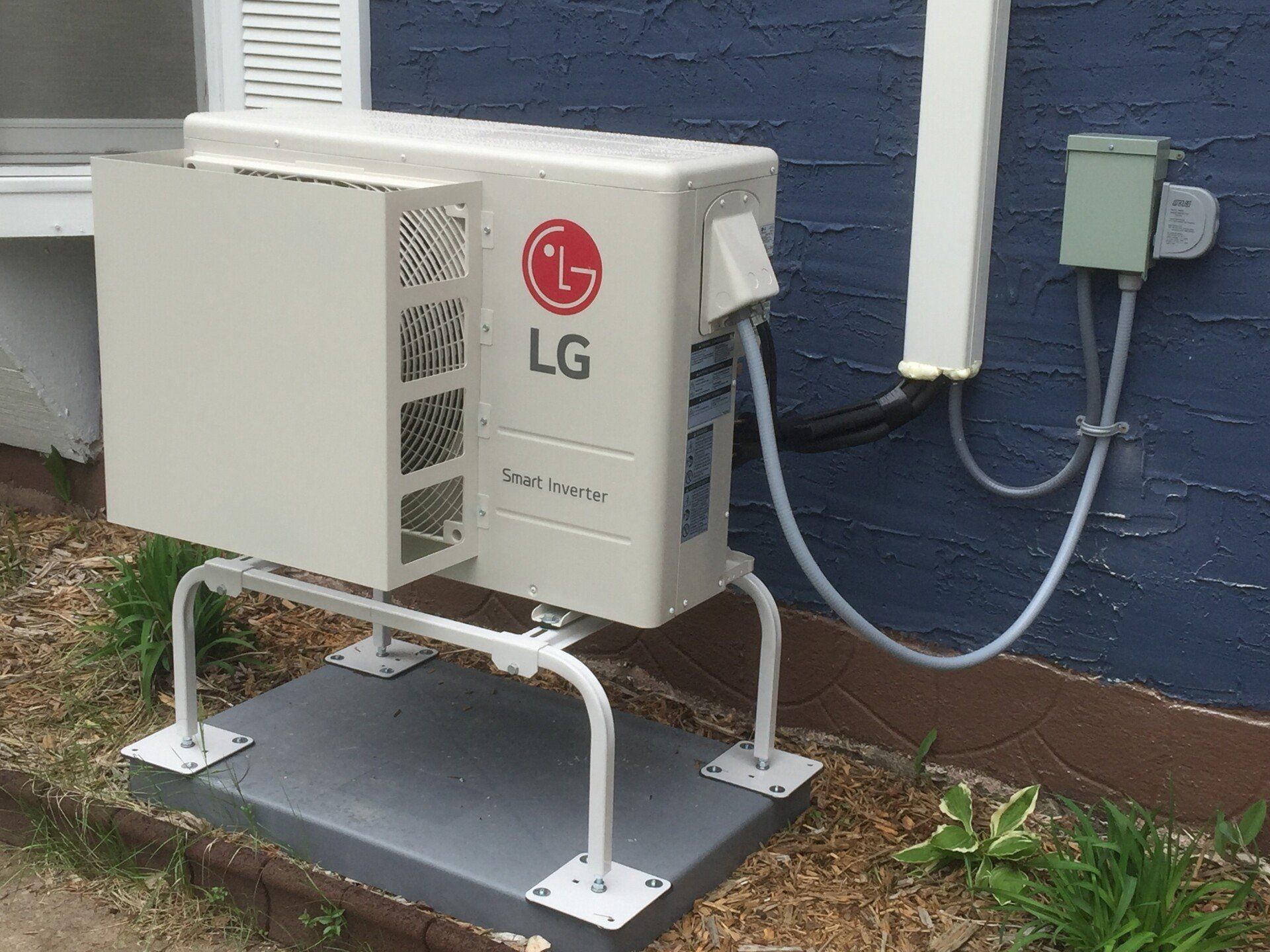 LG air conditioner unit mounted on a platform outside a blue house; electrical box and conduit are also visible.