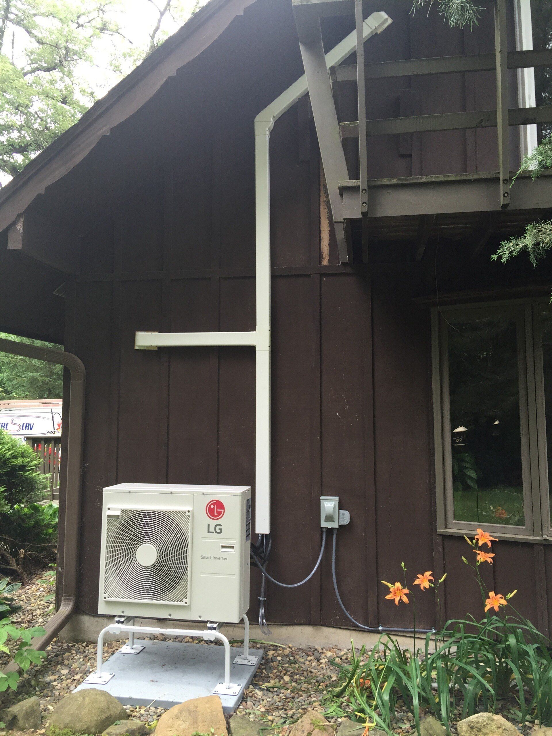 An LG air conditioning unit is mounted outside a brown building. White pipes run up the wall, near a wooden deck.