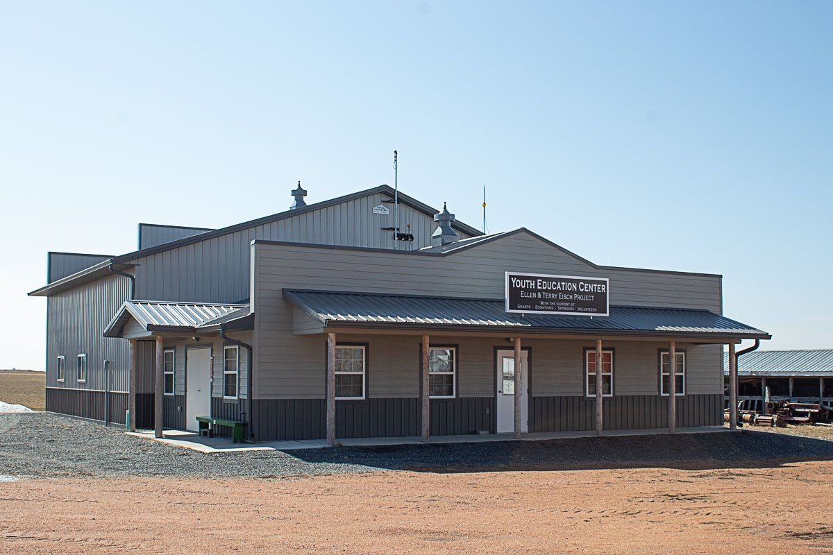 A tan and gray metal building with a covered porch and sign in a rural setting under a clear sky.