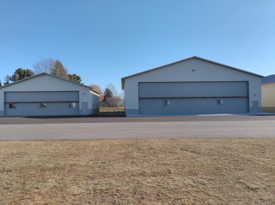 Two light-colored airplane hangars with large gray doors sit next to each other on a paved area, under a blue sky.