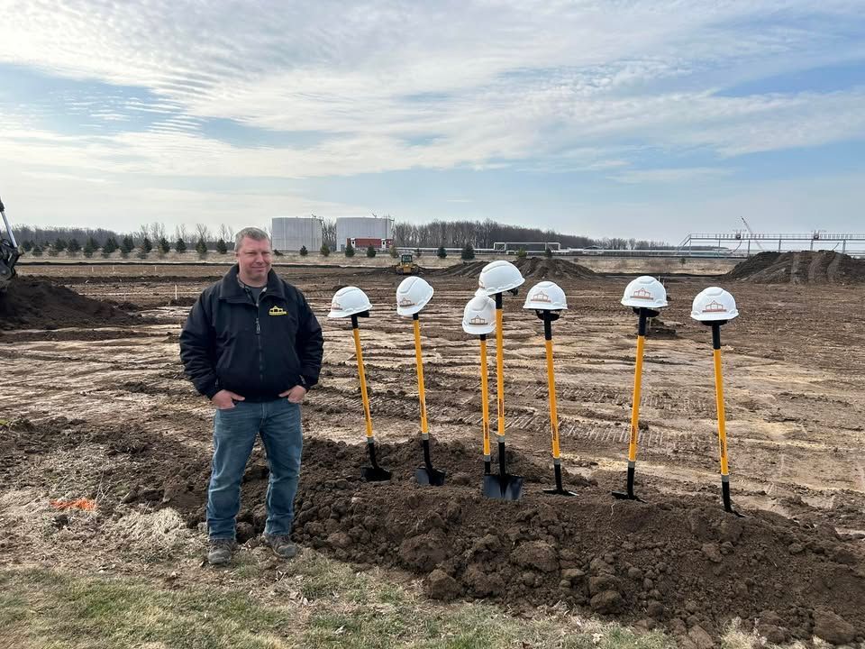 Man standing in front of shovels with hard hats, at a groundbreaking ceremony on a dirt field.