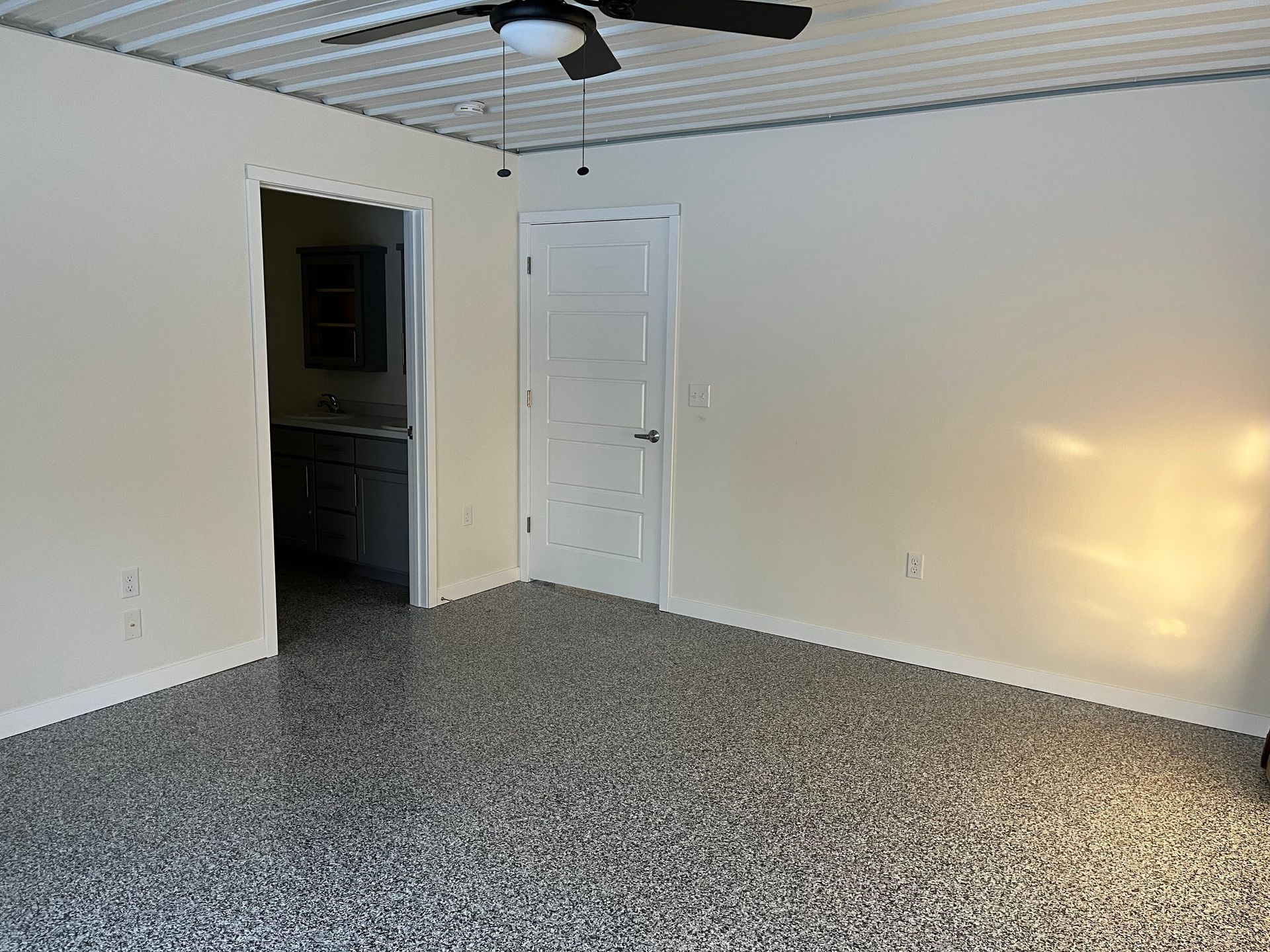 Interior view of a room with a gray speckled floor, door, and ceiling fan. Light-colored walls and a bathroom doorway.