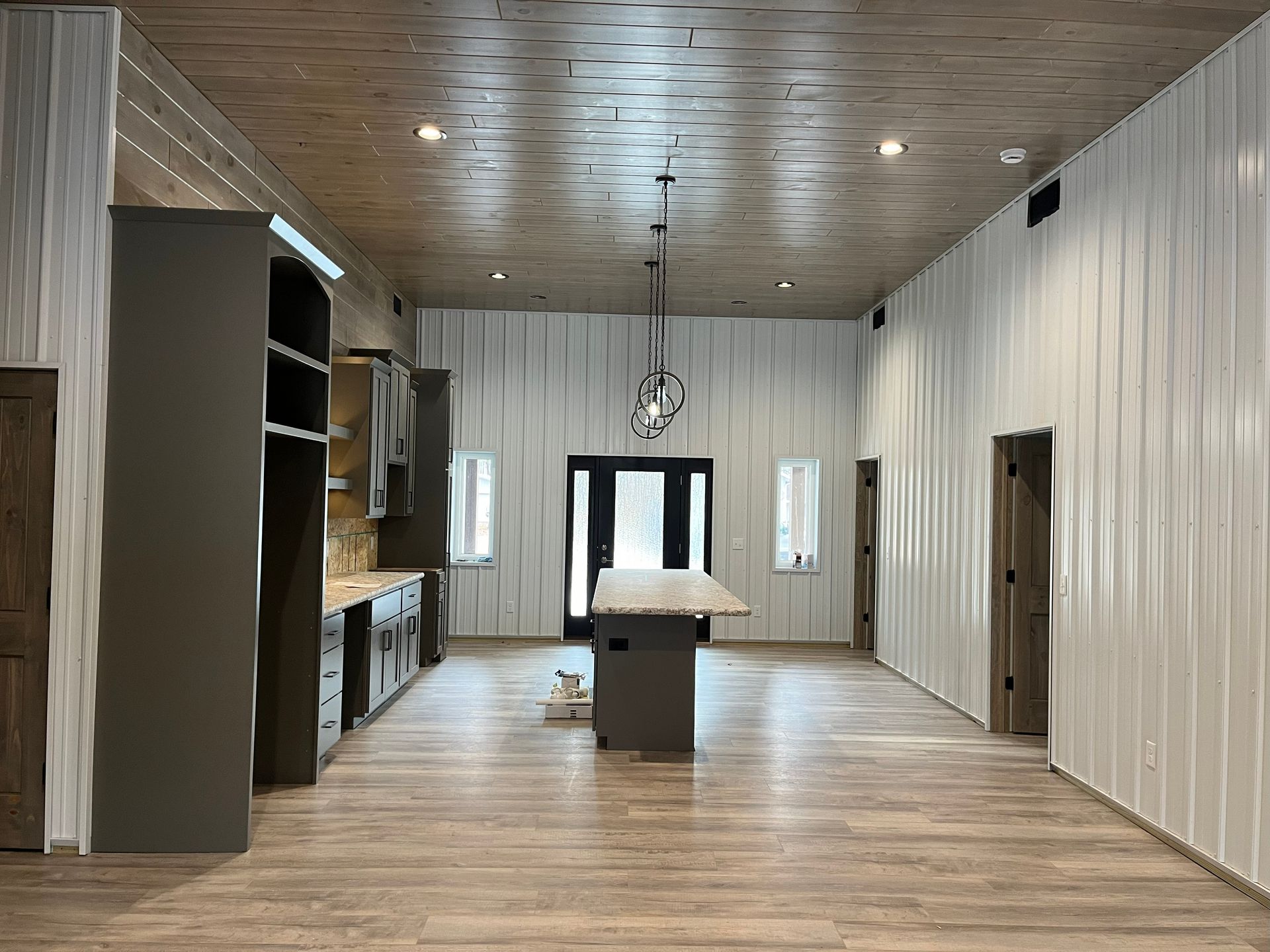 Spacious, modern interior with light wood floors, gray cabinets, and corrugated white walls. A black door is in the center.