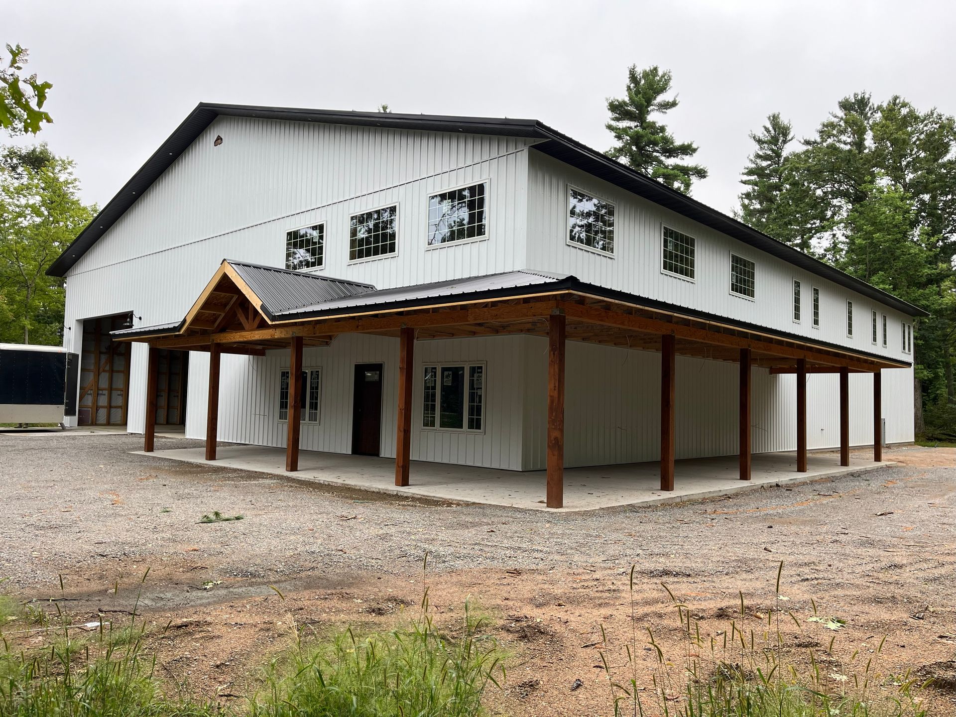 White two-story building with a porch, brown beams, and windows. Gravel ground, trees in the background.