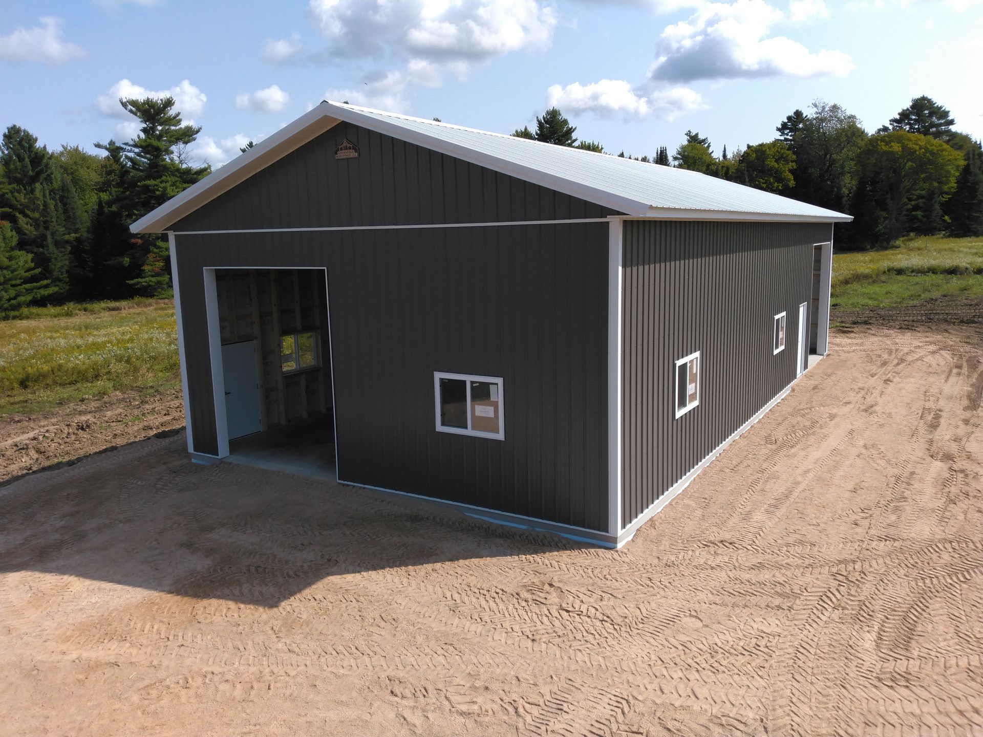 Dark green metal garage with white trim, open door, and two windows on a gravel lot.