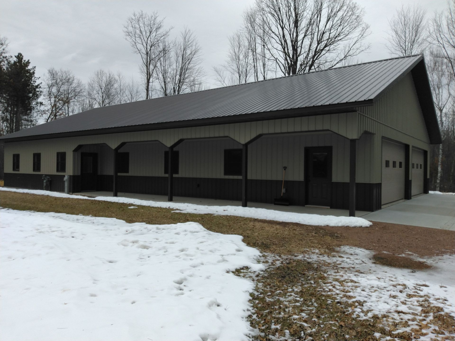 Long, tan and brown metal building with two garage doors and a covered porch, snow on the ground.