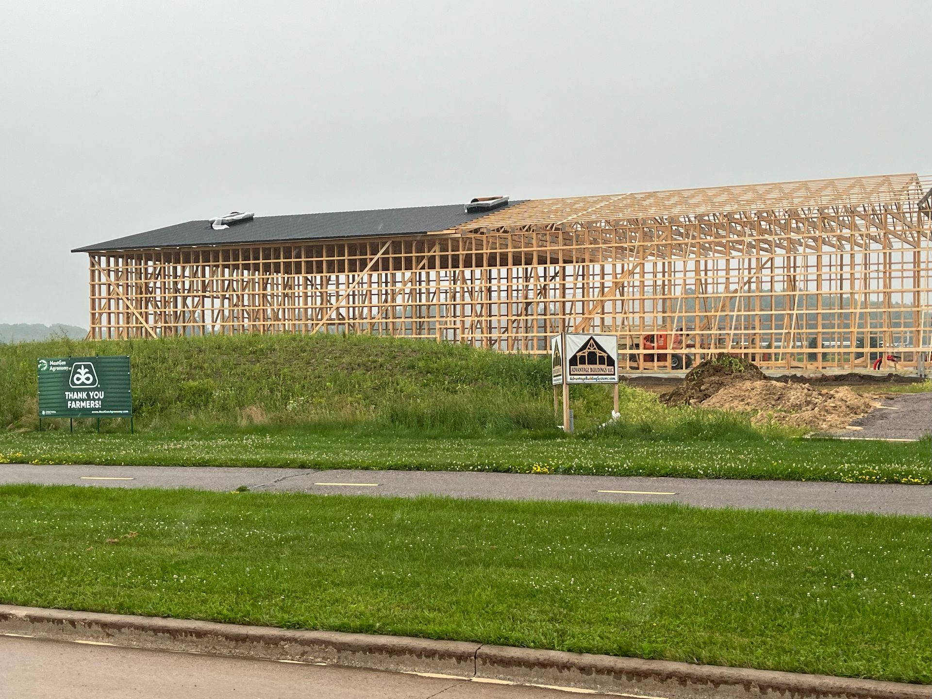 A large wooden building under construction with a partly finished roof. Green grass and signage in front.