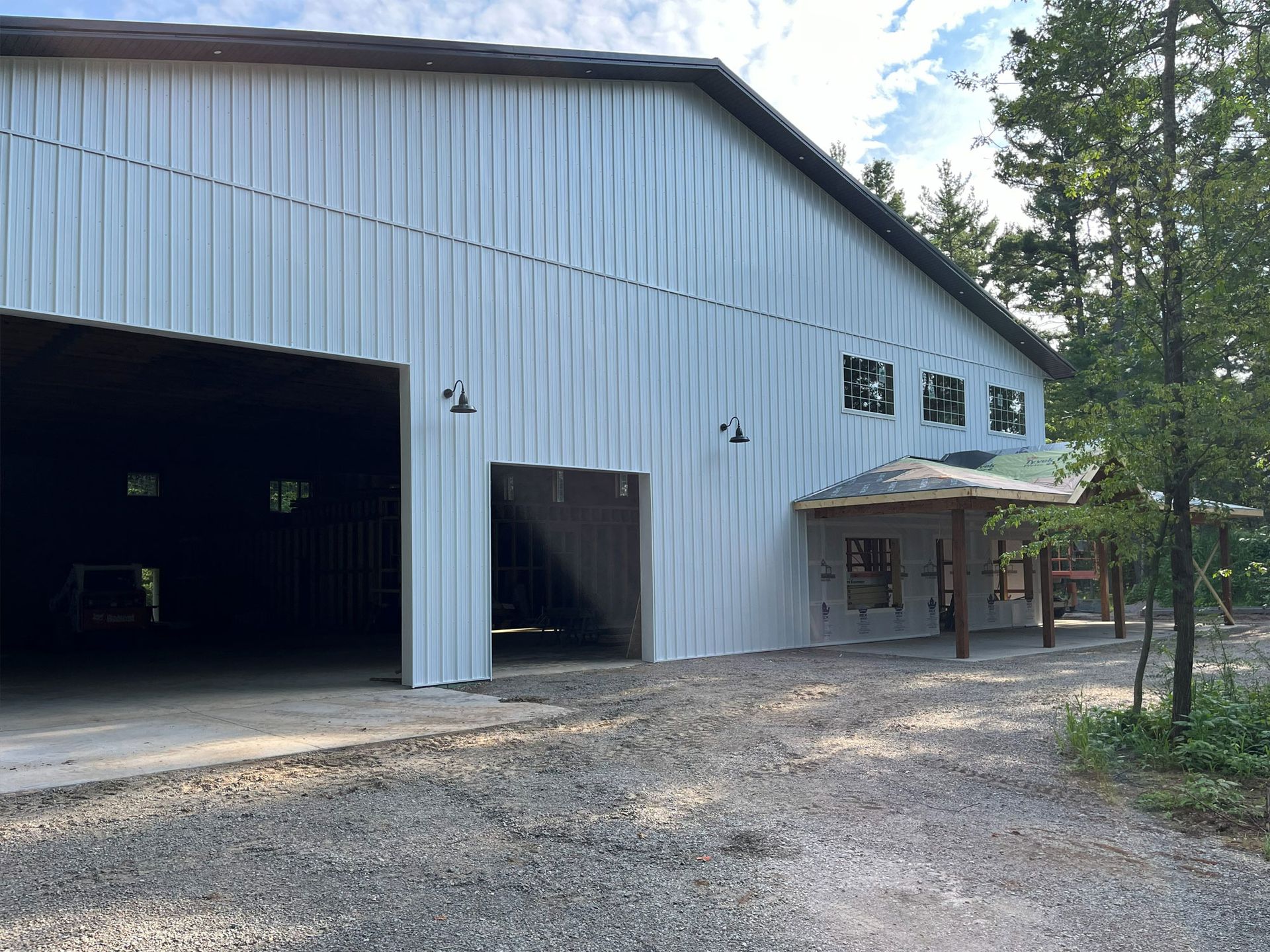 Large metal building with open bays, gravel driveway, and surrounding trees.