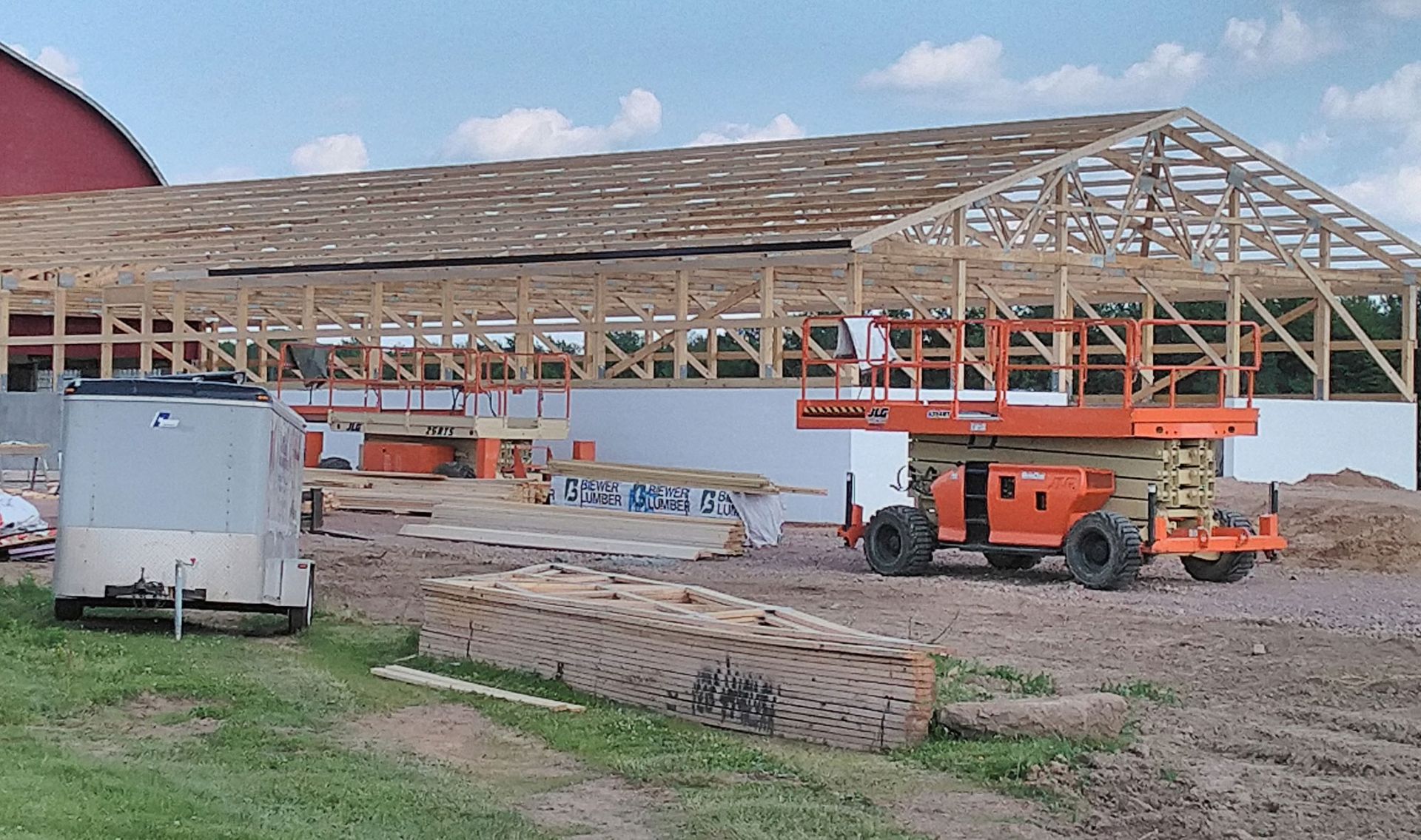 Construction of a wooden barn frame, orange lift, and white trailer on a grassy field.