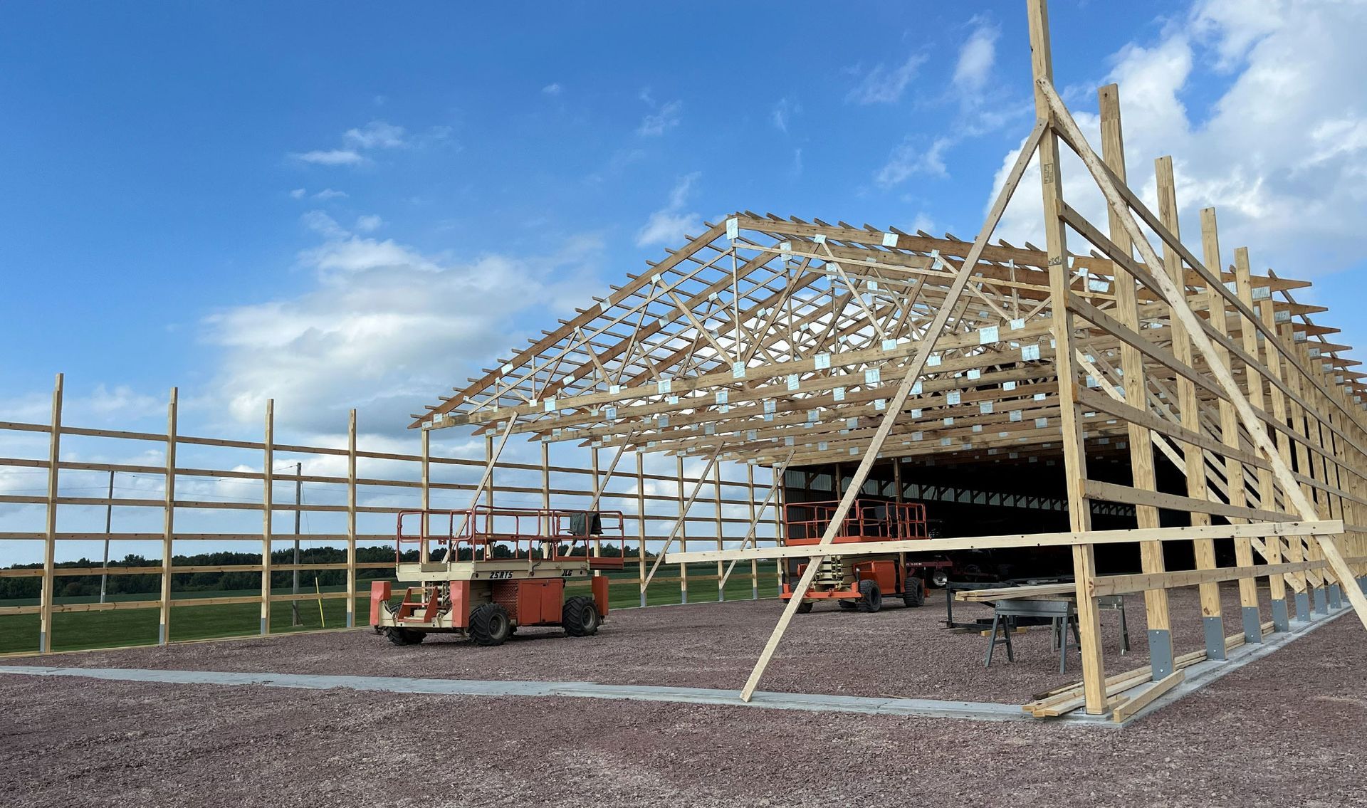 Barn under construction with wooden frame, equipment, and gravel base under a blue sky.