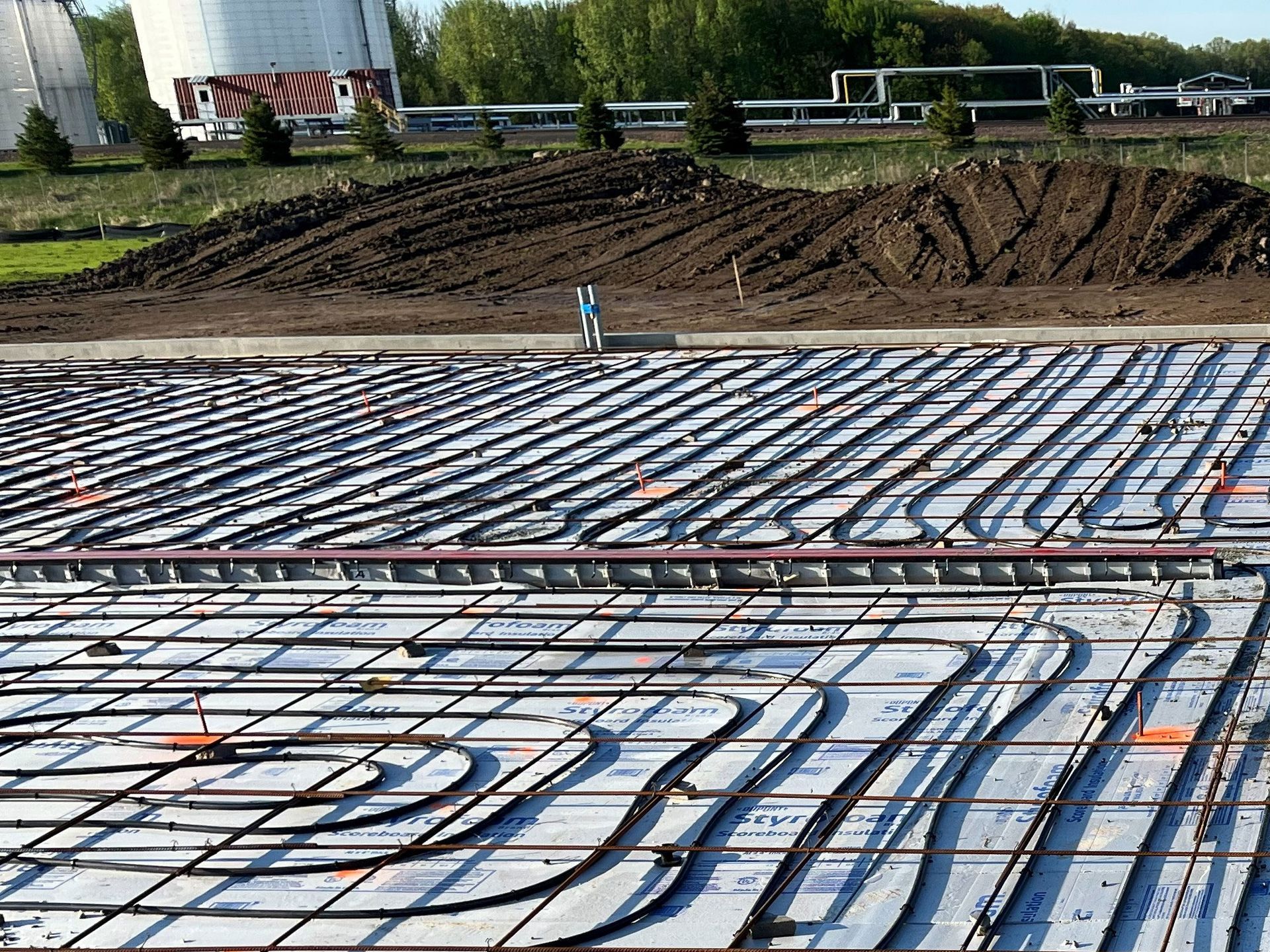 Piping system installation on a concrete foundation, likely for heating. Industrial setting, daytime.