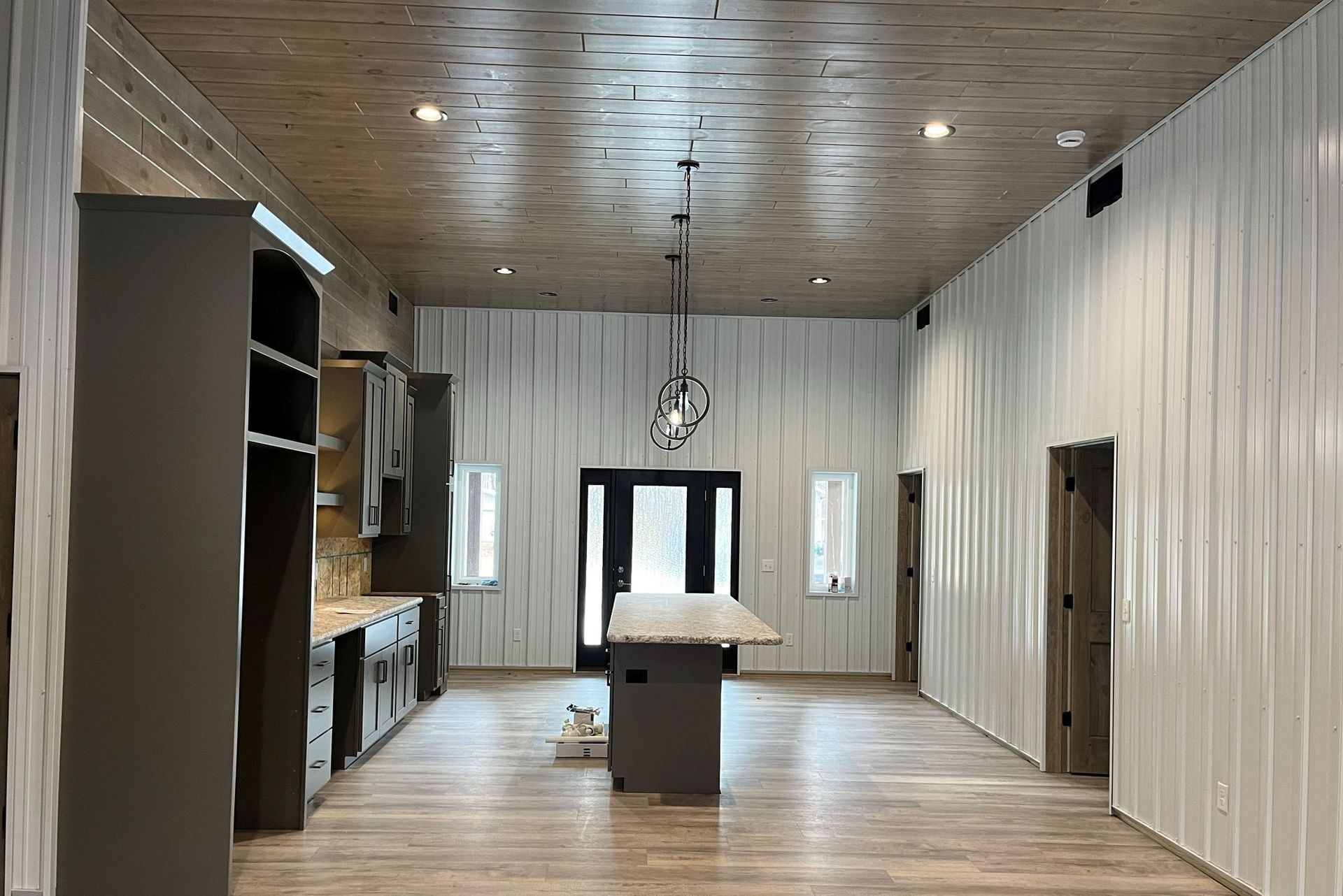 Interior of a large, open space with a kitchen area and central island. White corrugated walls, wood ceiling and flooring.