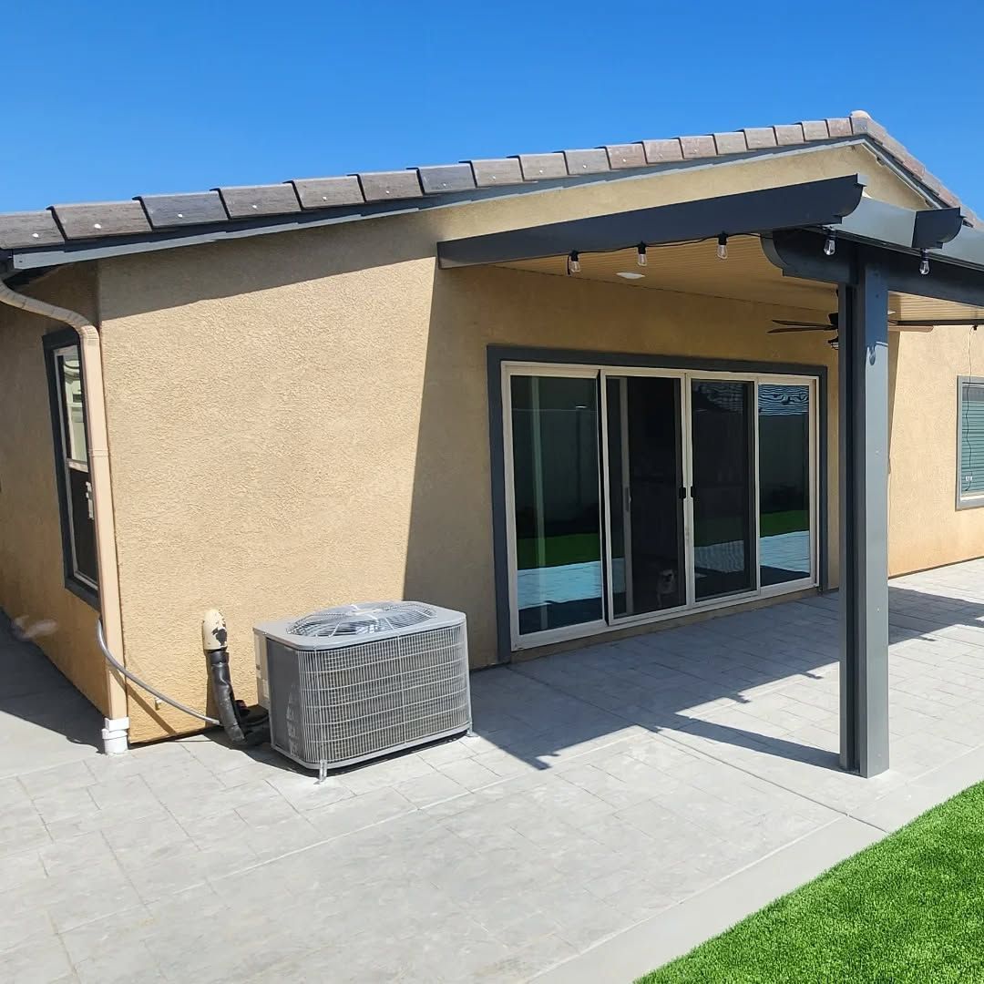 A beige stucco house with a tiled roof, patio door, and attached pergola over a concrete patio with an AC unit.