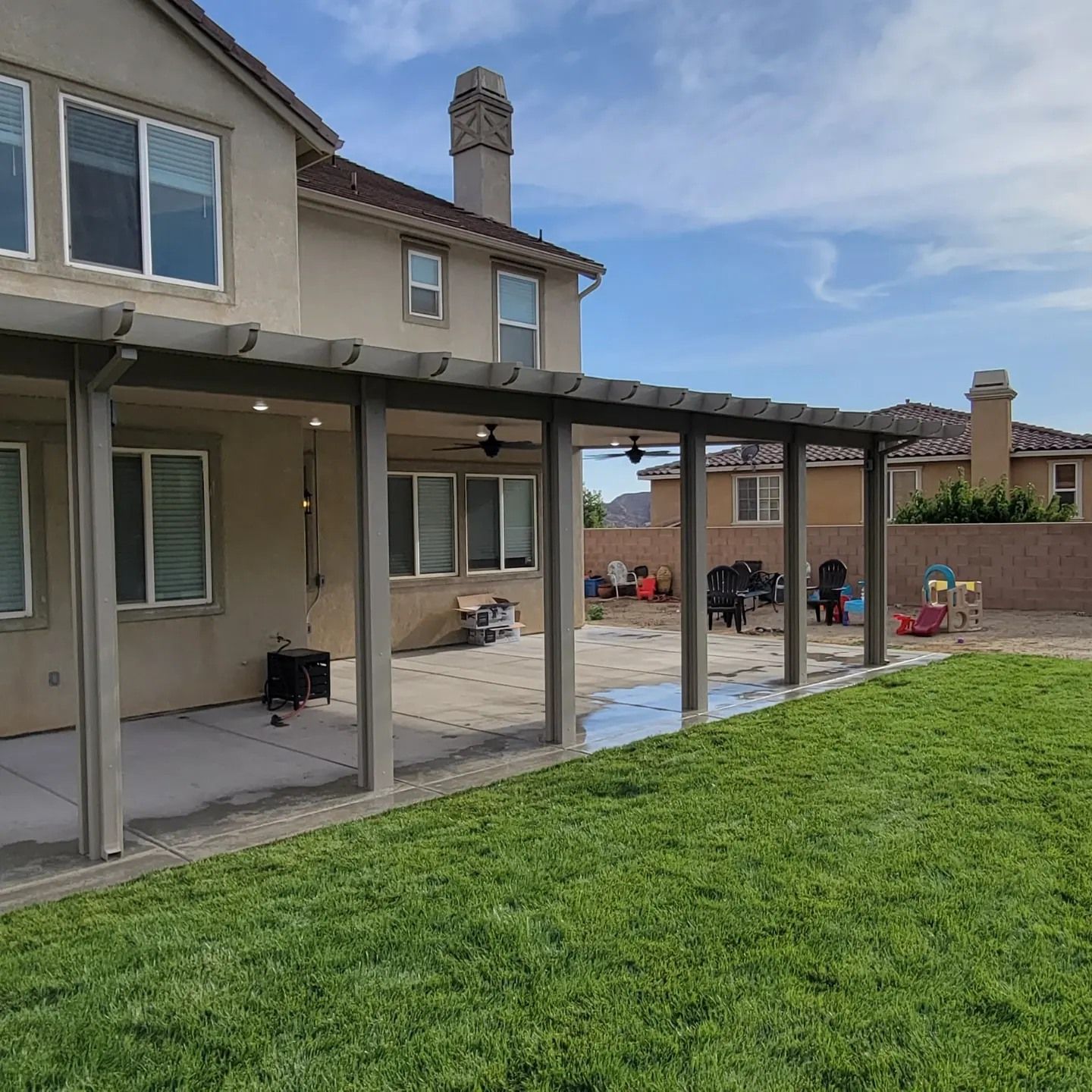 A beige two-story house with a large wooden patio cover extending over a concrete slab and green grass lawn.