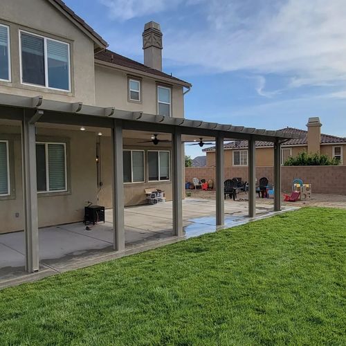 A beige two-story house with a large wooden patio cover extending over a concrete slab and green grass lawn.