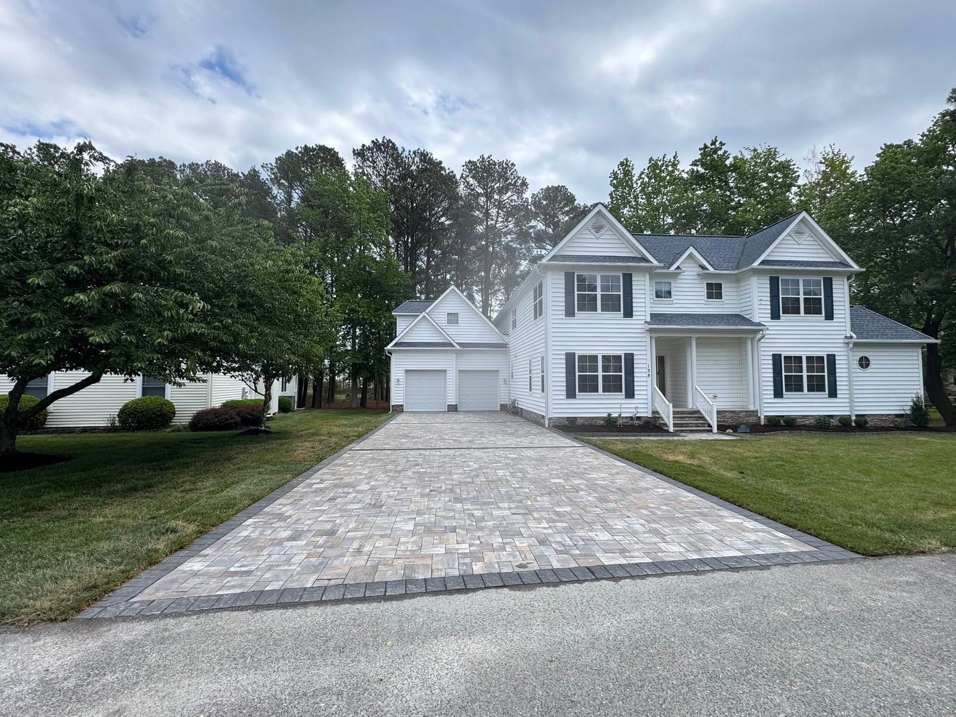 White two-story house with a stone driveway, detached garage, and large trees under cloudy skies