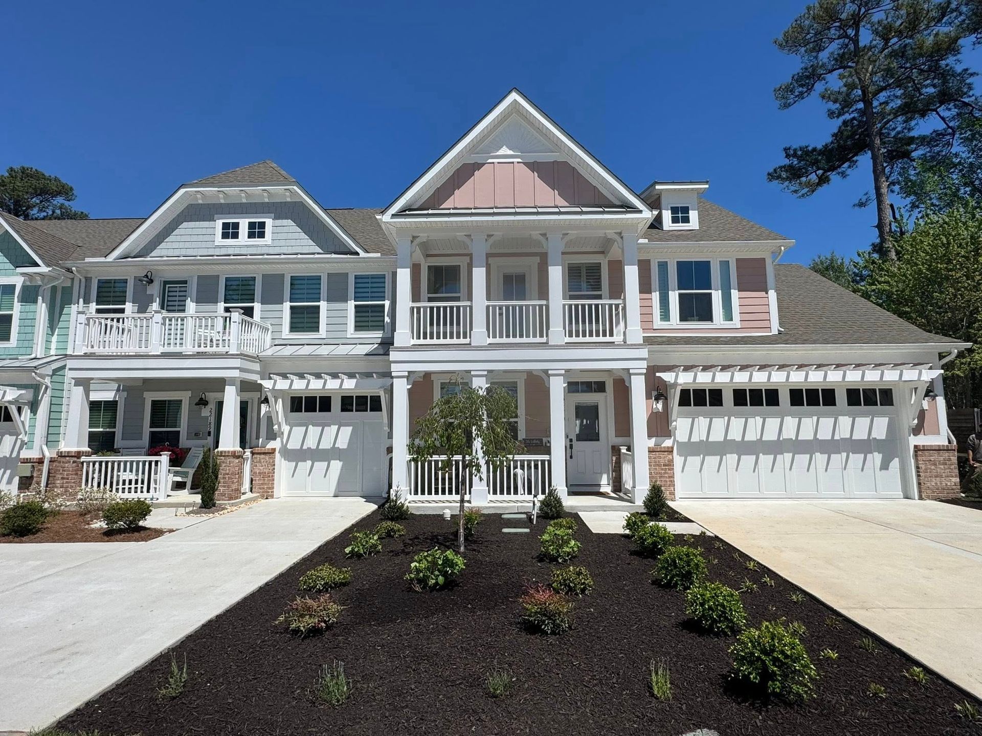 Large white coastal-style house with multiple gables, garages, and a freshly landscaped front yard under a clear blue sky