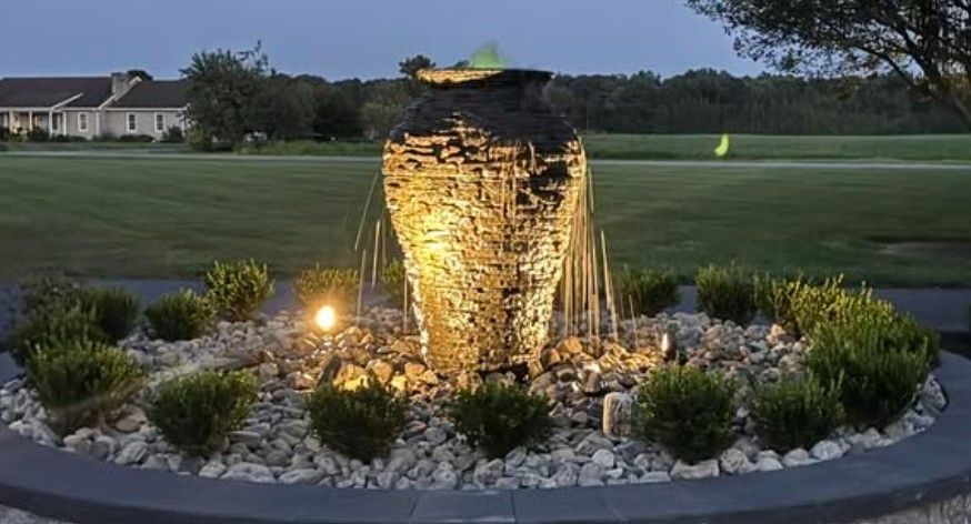 Stone fountain illuminated at dusk, surrounded by shrubs and rocks.