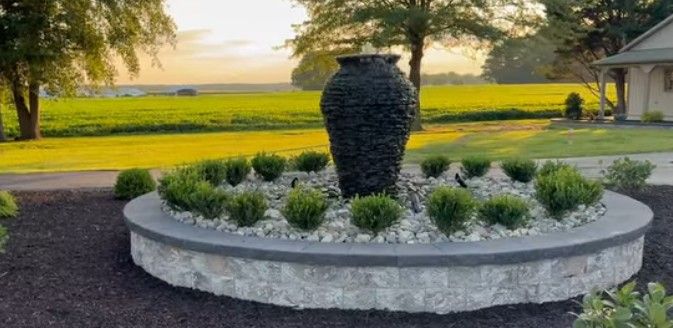 A decorative stone fountain with a large black urn at its center. Green shrubs and white stones encircle the fountain.
