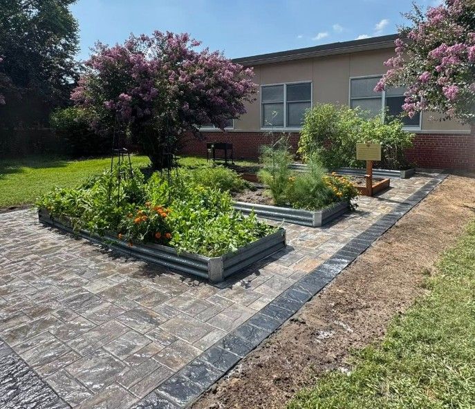 Raised garden beds with flourishing plants on a brick patio. Building in the background.