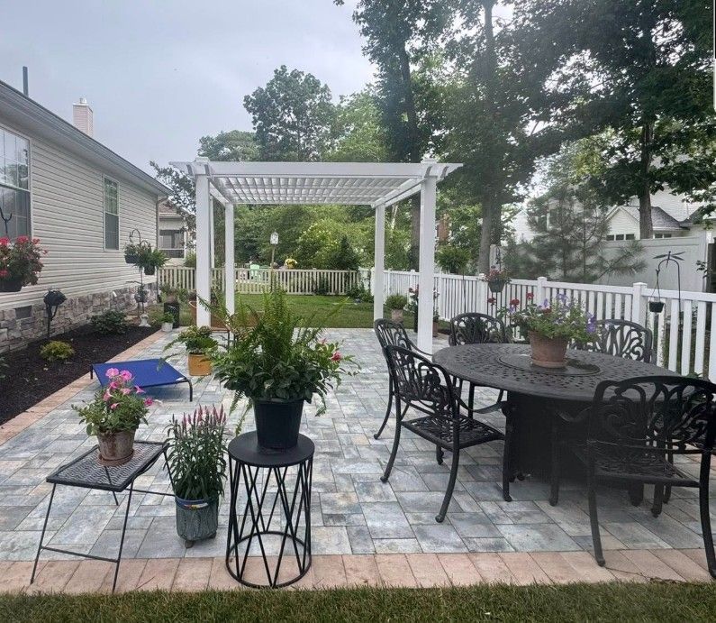 Backyard patio with patterned pavers, white pergola, and wrought-iron furniture.