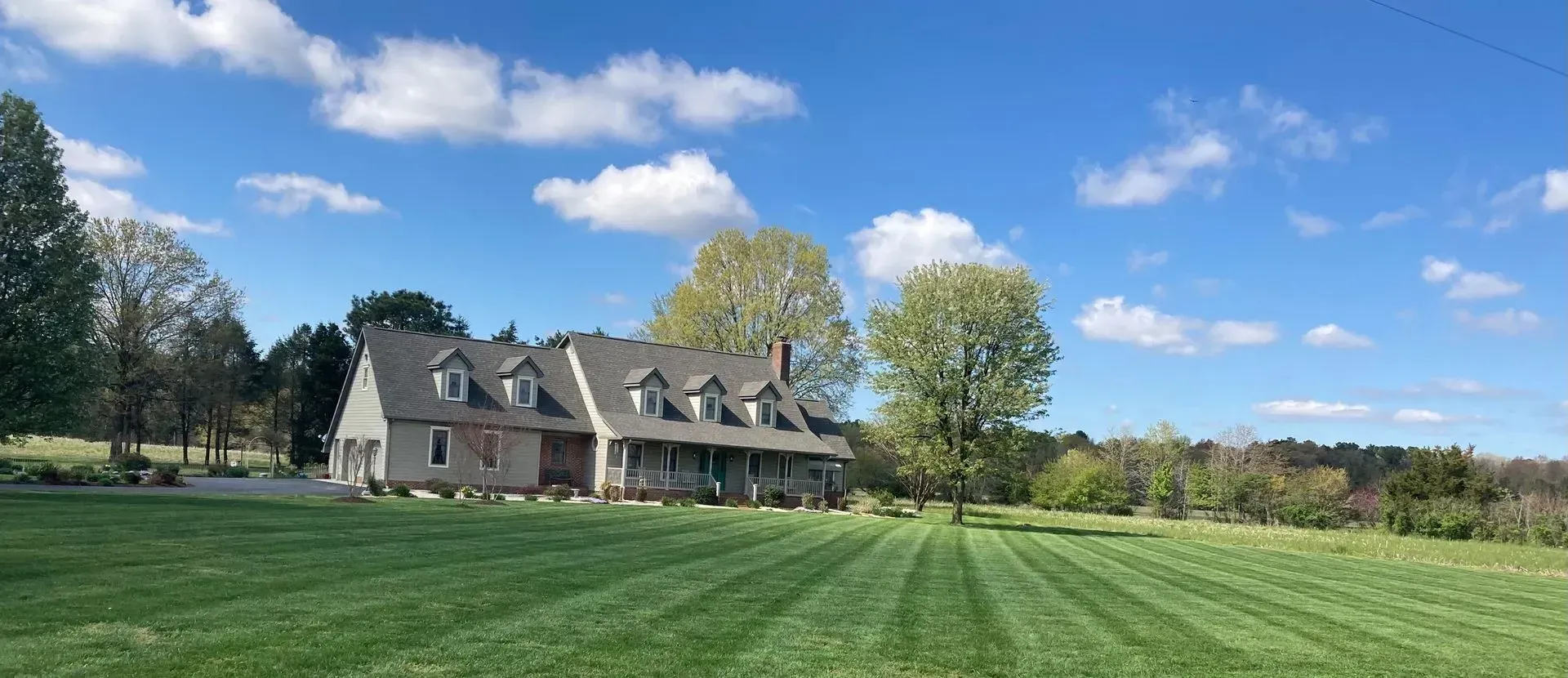 Large stone house with dormer windows beside a green field under a blue sky with clouds