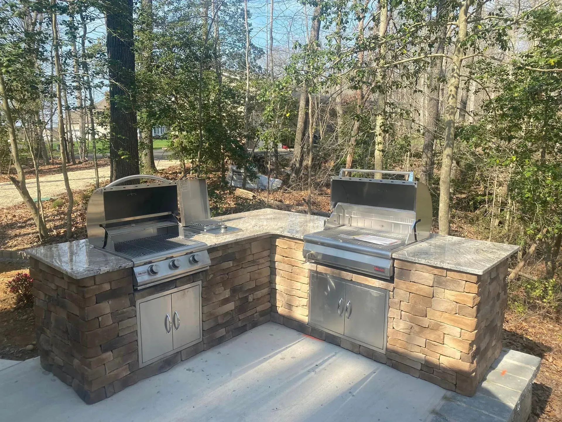 Outdoor stone barbecue kitchen with two stainless steel grills on a patio in a wooded yard