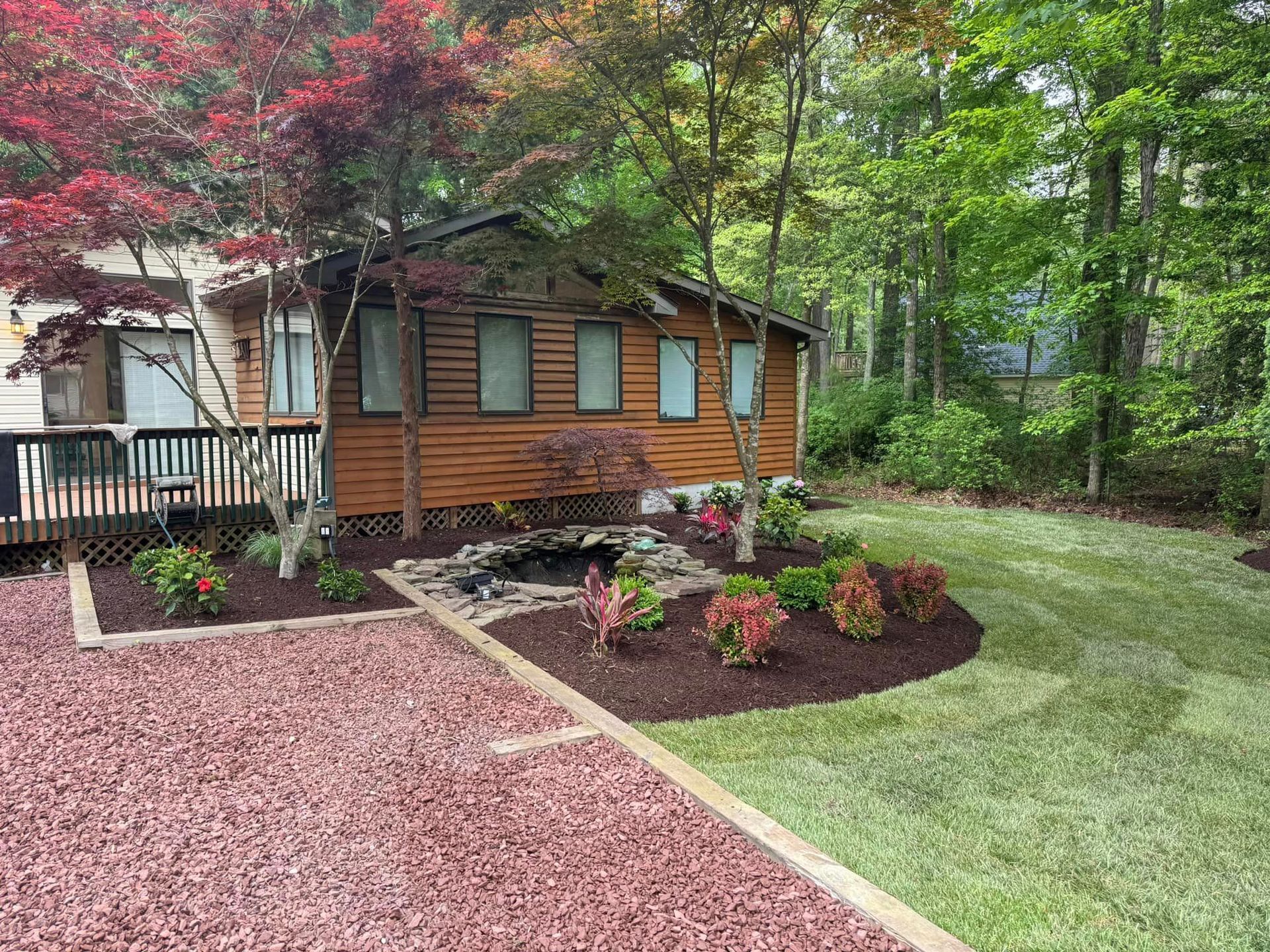 A cabin with wooden siding, a small pond, and a manicured garden. Surrounded by trees and mulch.