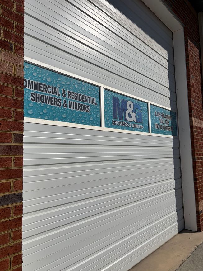 A white garage door features three window signs advertising M&G Showers & Mirrors, framed by brick walls.