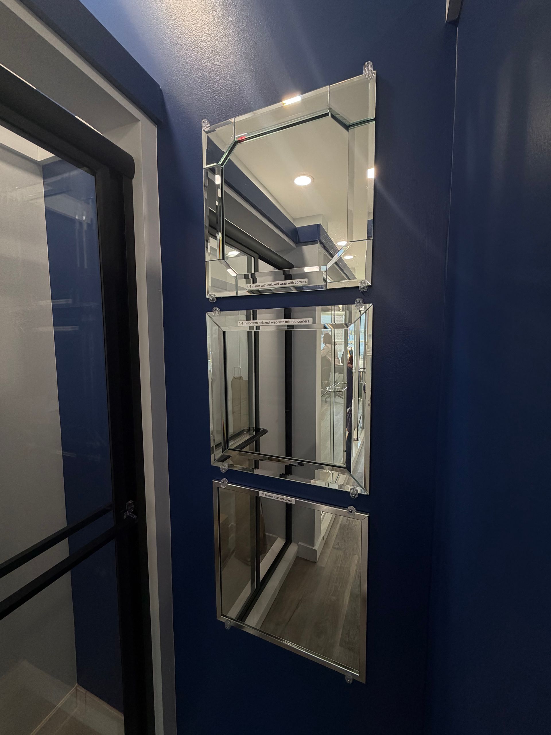 Three rectangular, beveled-edge mirrors are stacked vertically on a dark blue wall next to a glass-paneled doorway.