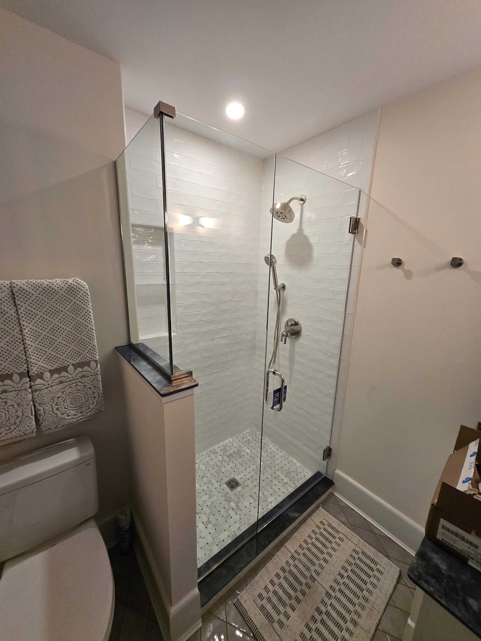 A walk-in shower with white tiled walls, a glass enclosure, a handheld showerhead, and black stone trim in a bathroom.