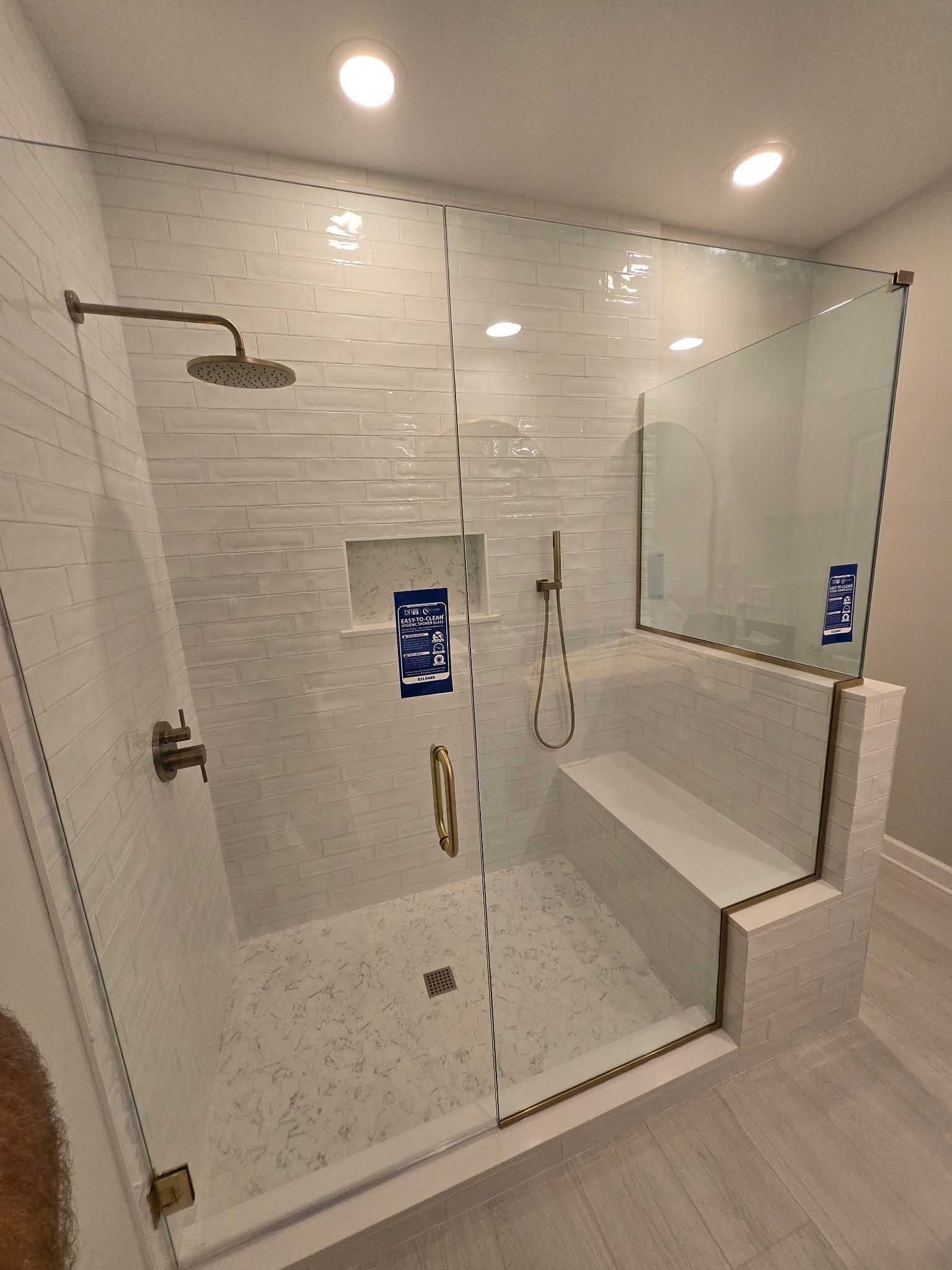 A modern tiled walk-in shower with a glass enclosure, built-in bench, overhead rainfall showerhead, and gold hardware.