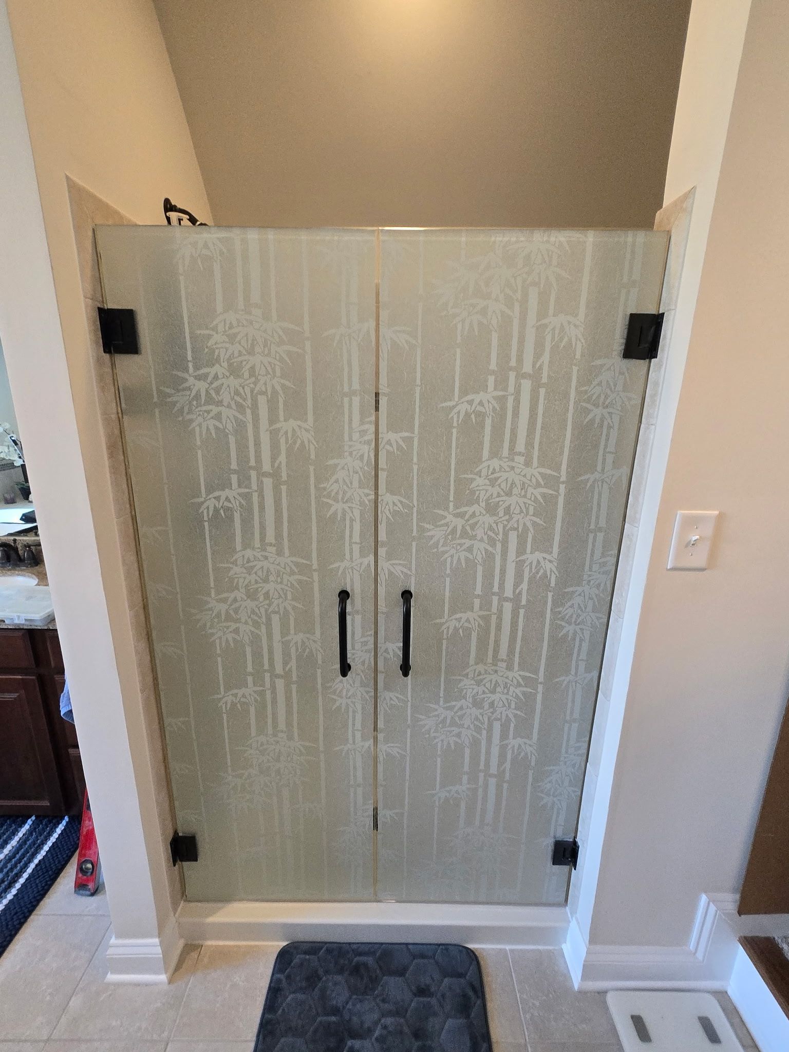 A glass shower door with a frosted bamboo pattern, black hardware, and two handles installed in a beige bathroom.