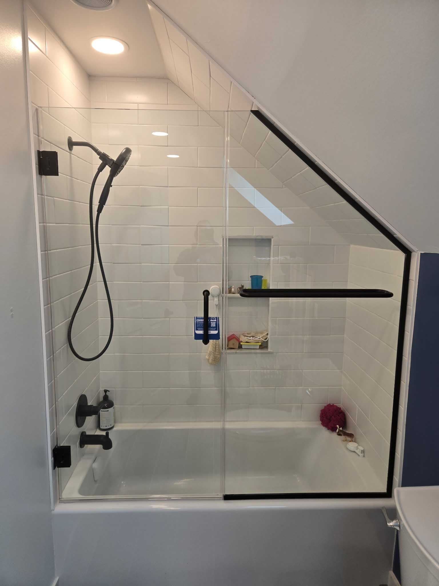 A white tiled bathtub and shower with a slanted glass enclosure, black hardware, and a built-in recessed niche.