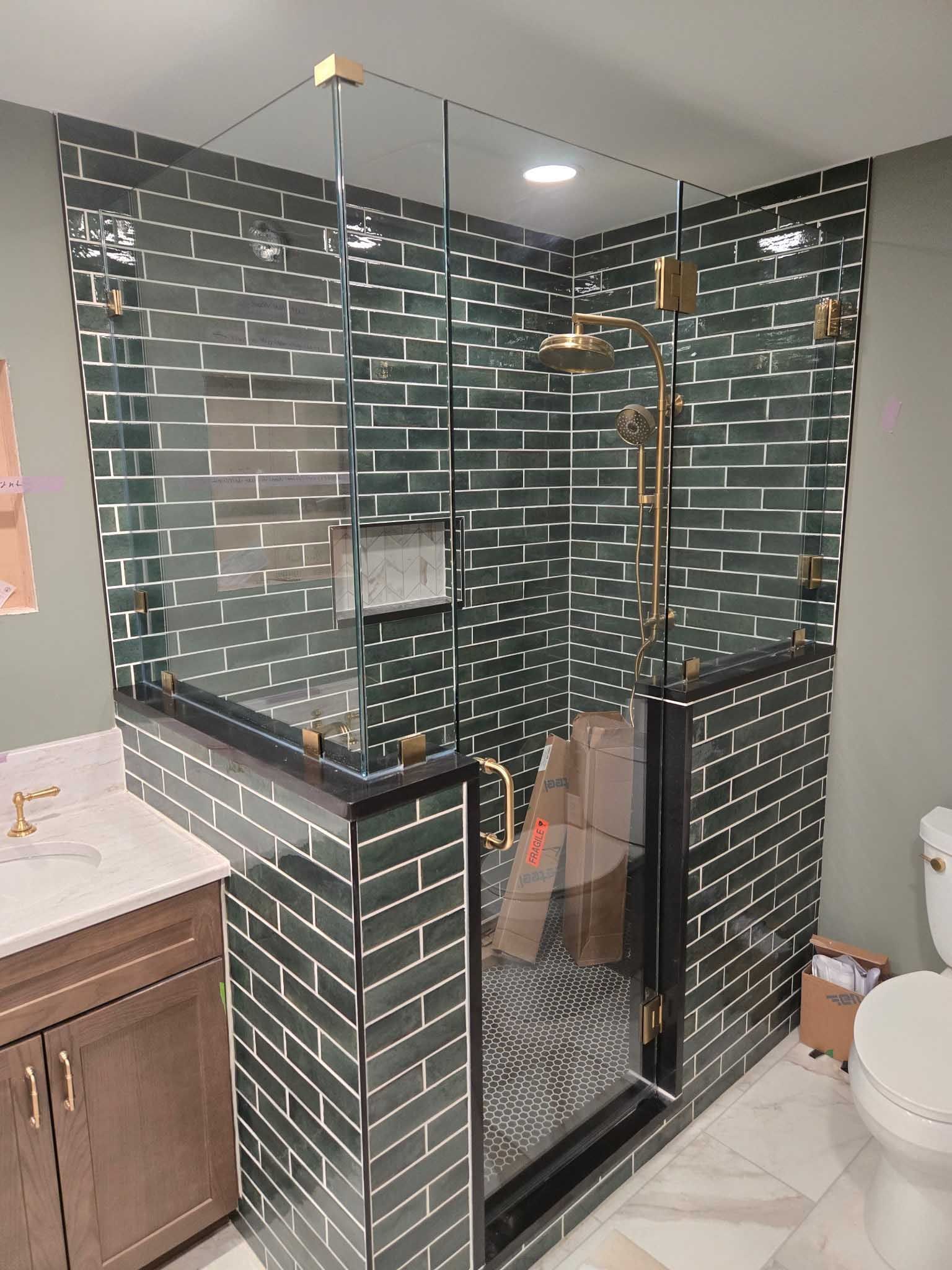A corner shower with dark green brick-patterned tiles, glass enclosure, gold fixtures, and a seated bench inside.