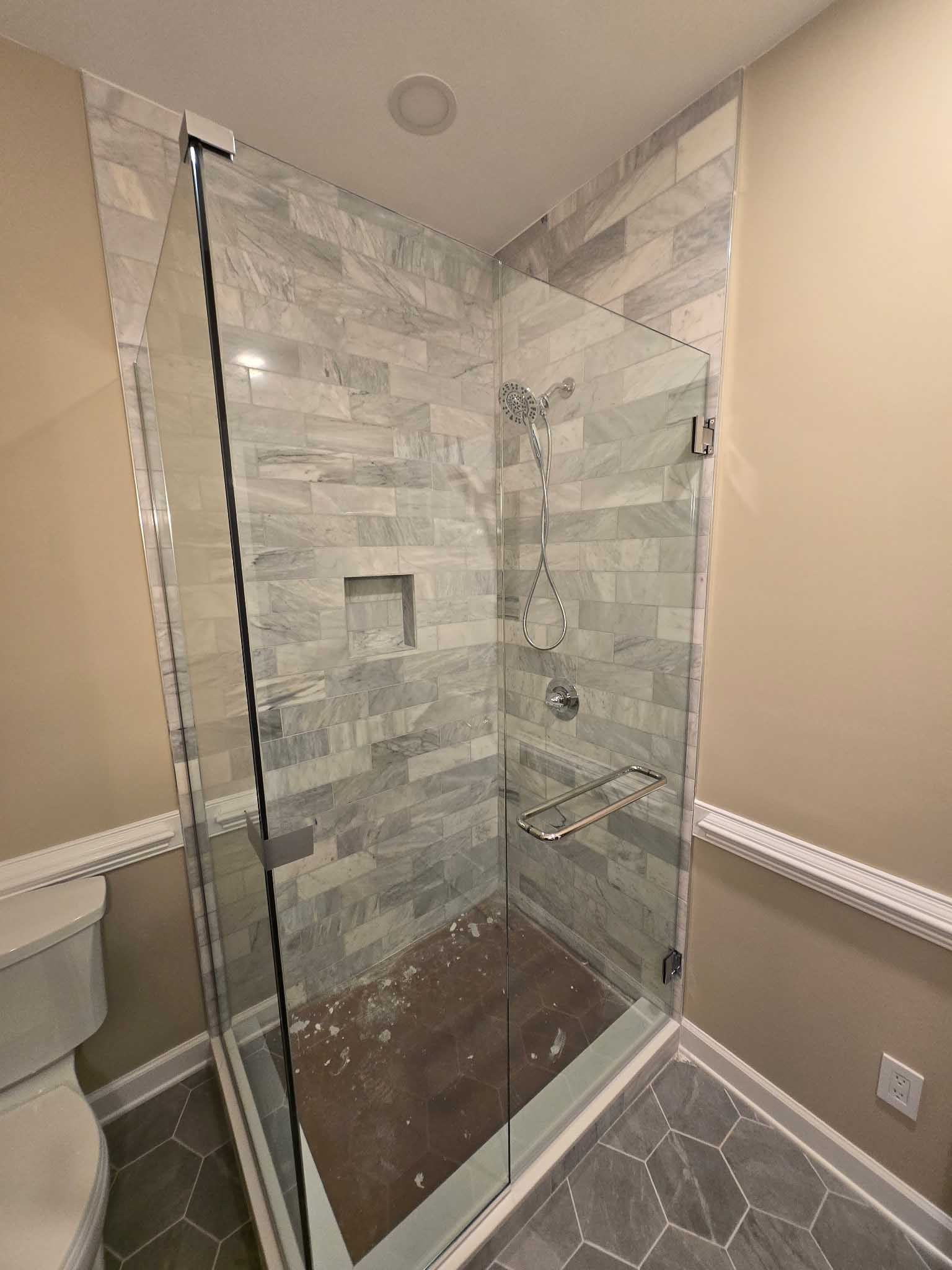 A glass-enclosed shower with gray stacked stone walls and a dark shower floor, set in a bathroom with hexagonal tiles.