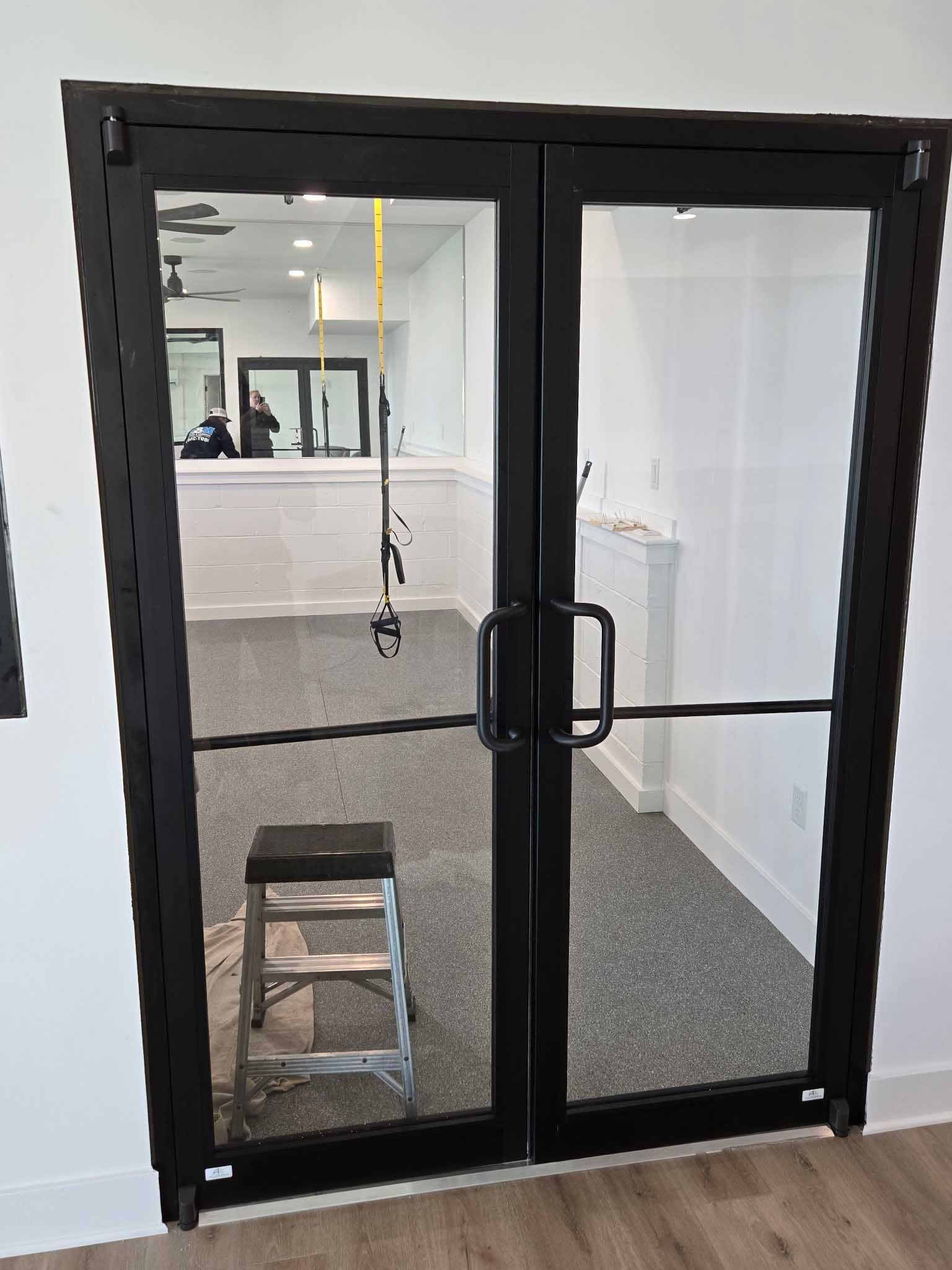 Black double glass doors with a rectangular frame and metal handles, showing a room with carpet and a small metal step stool.