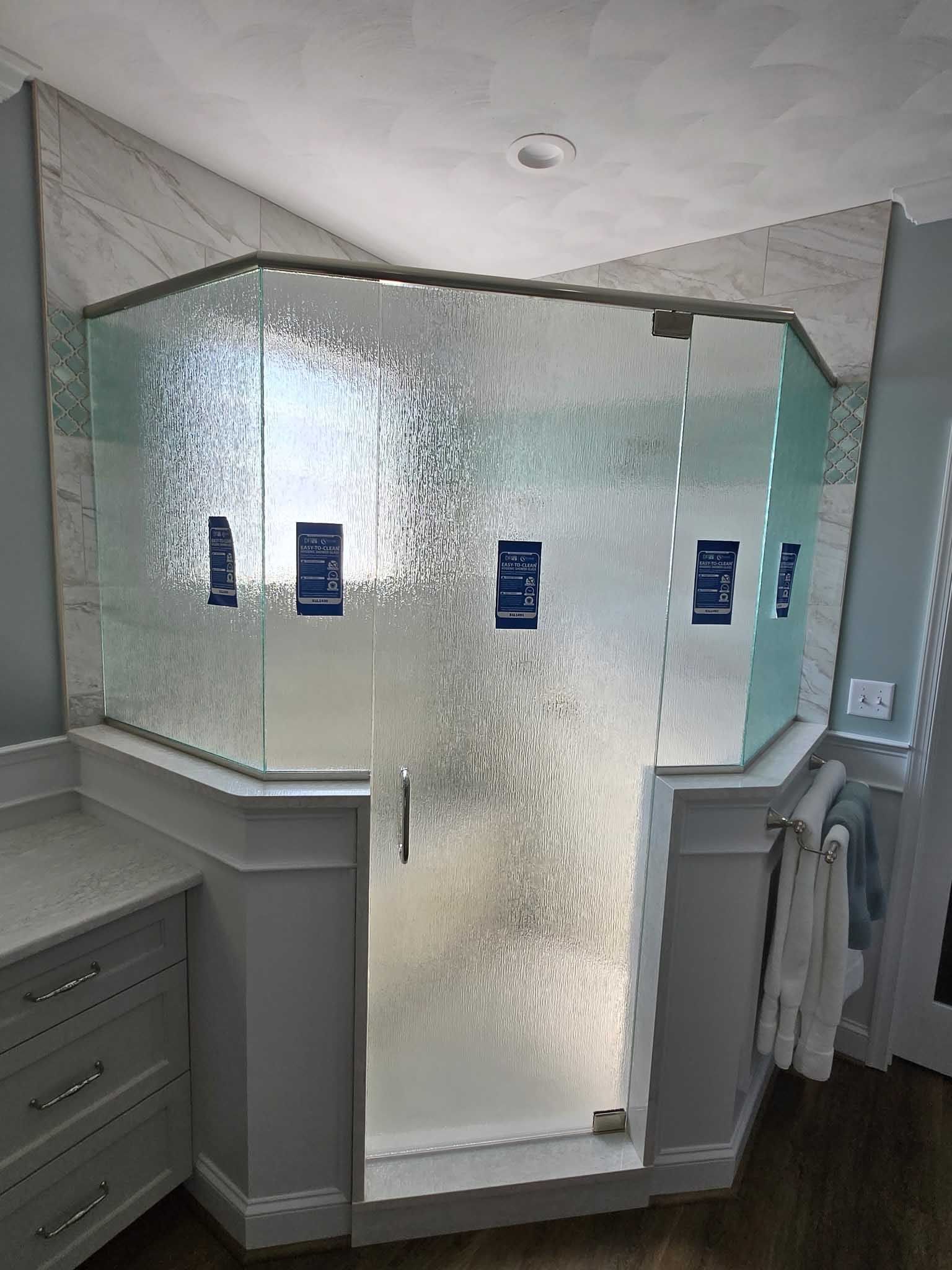 A neo-angle glass shower enclosure with textured privacy glass, installed in a bathroom corner with white wainscoting.