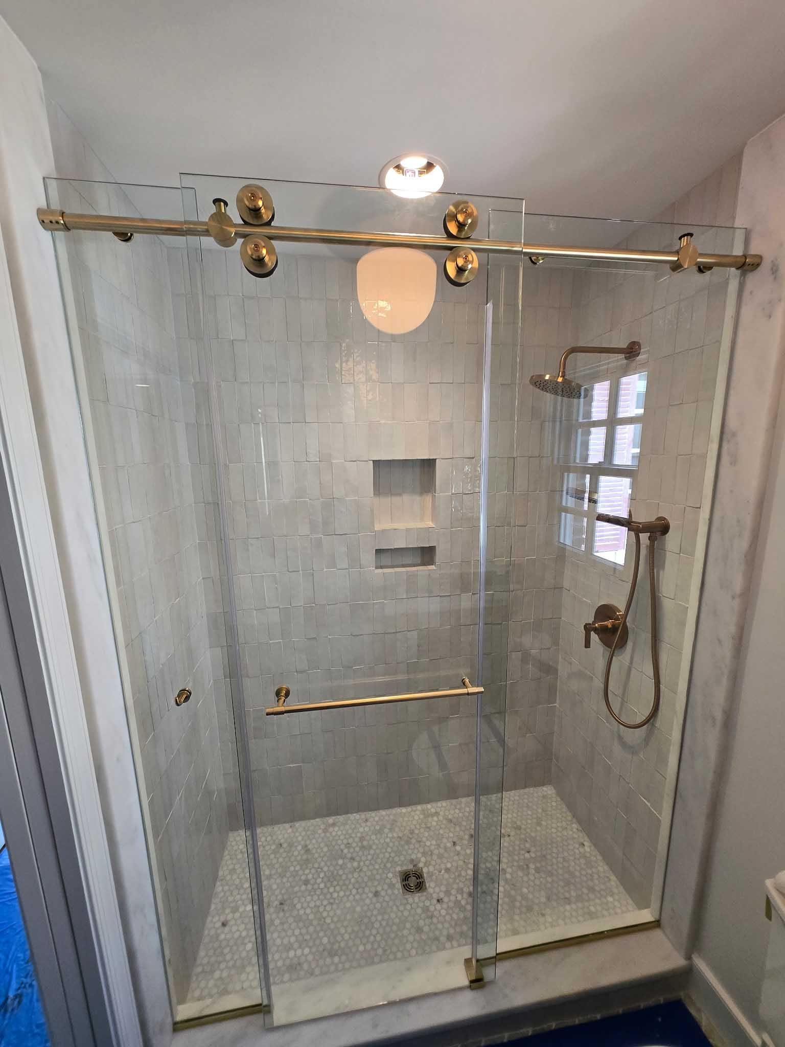 A glass walk-in shower with gold hardware, white marble tiled walls, a small built-in shelf, and a mosaic tiled floor.
