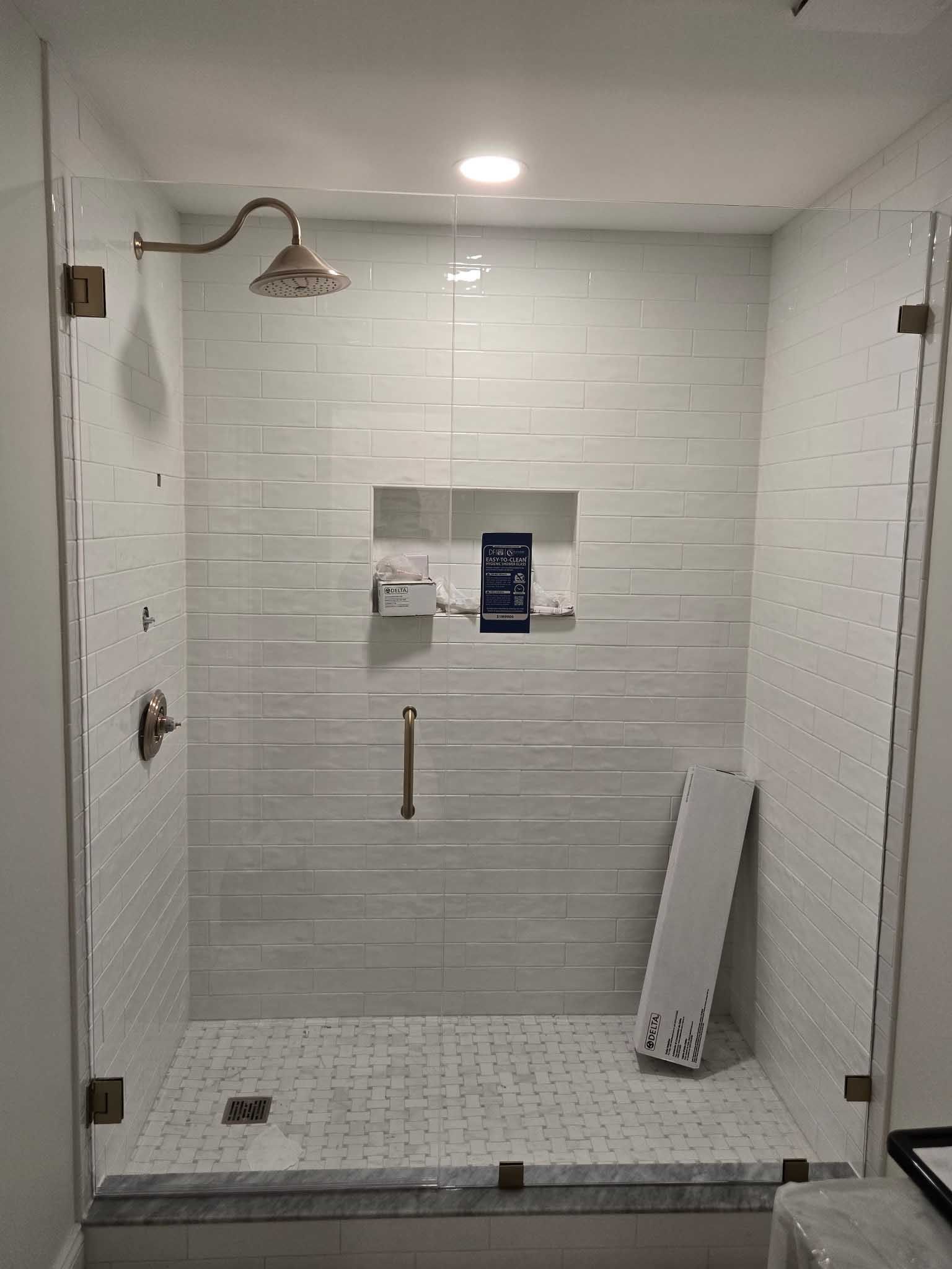 A walk-in shower with white tiled walls, a small recessed shelf, a rain showerhead, a metal grab bar, and a glass door.
