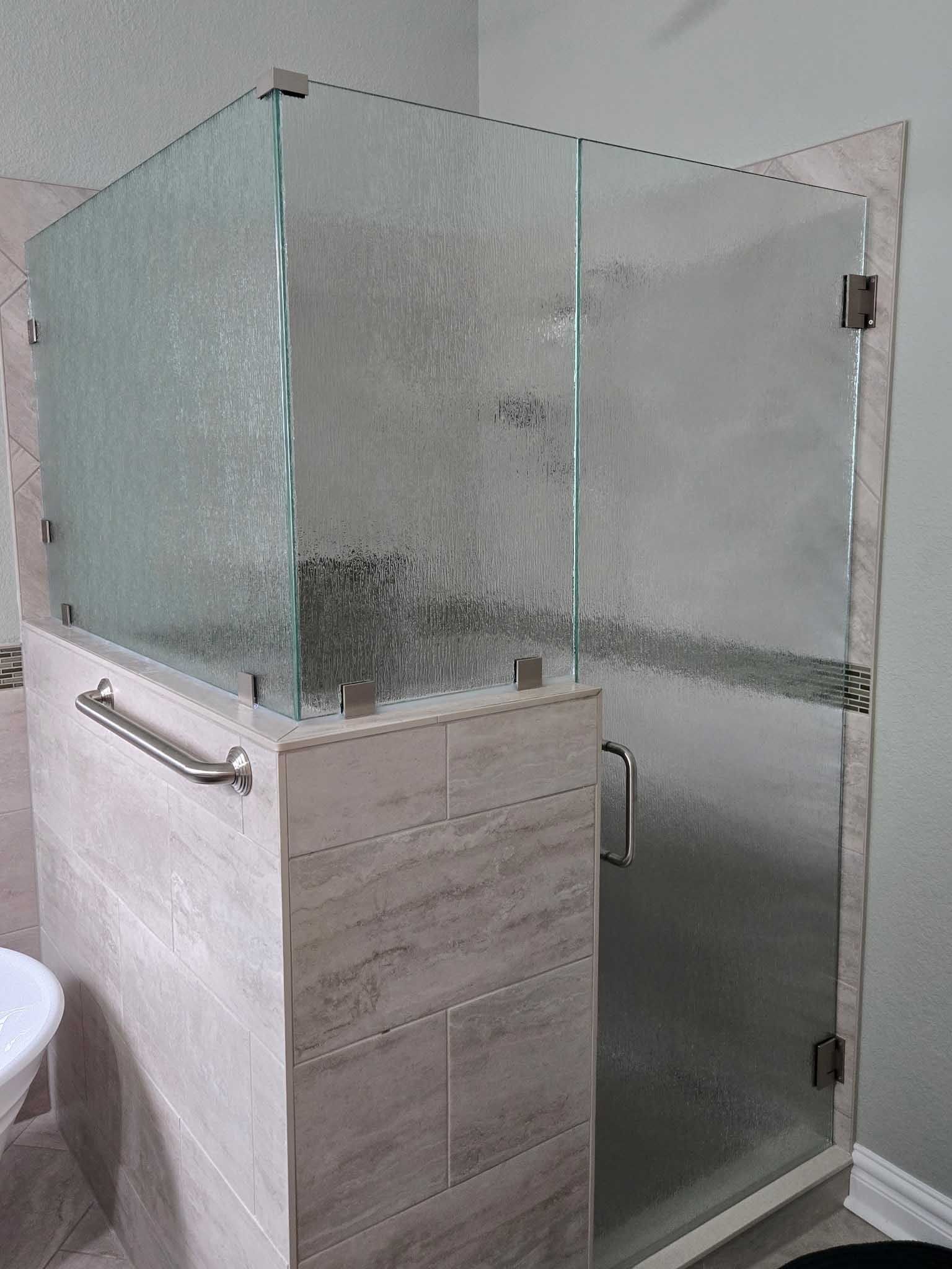 A glass shower enclosure with textured glass panels, a metal handle, and a grab bar installed on a tiled knee wall.