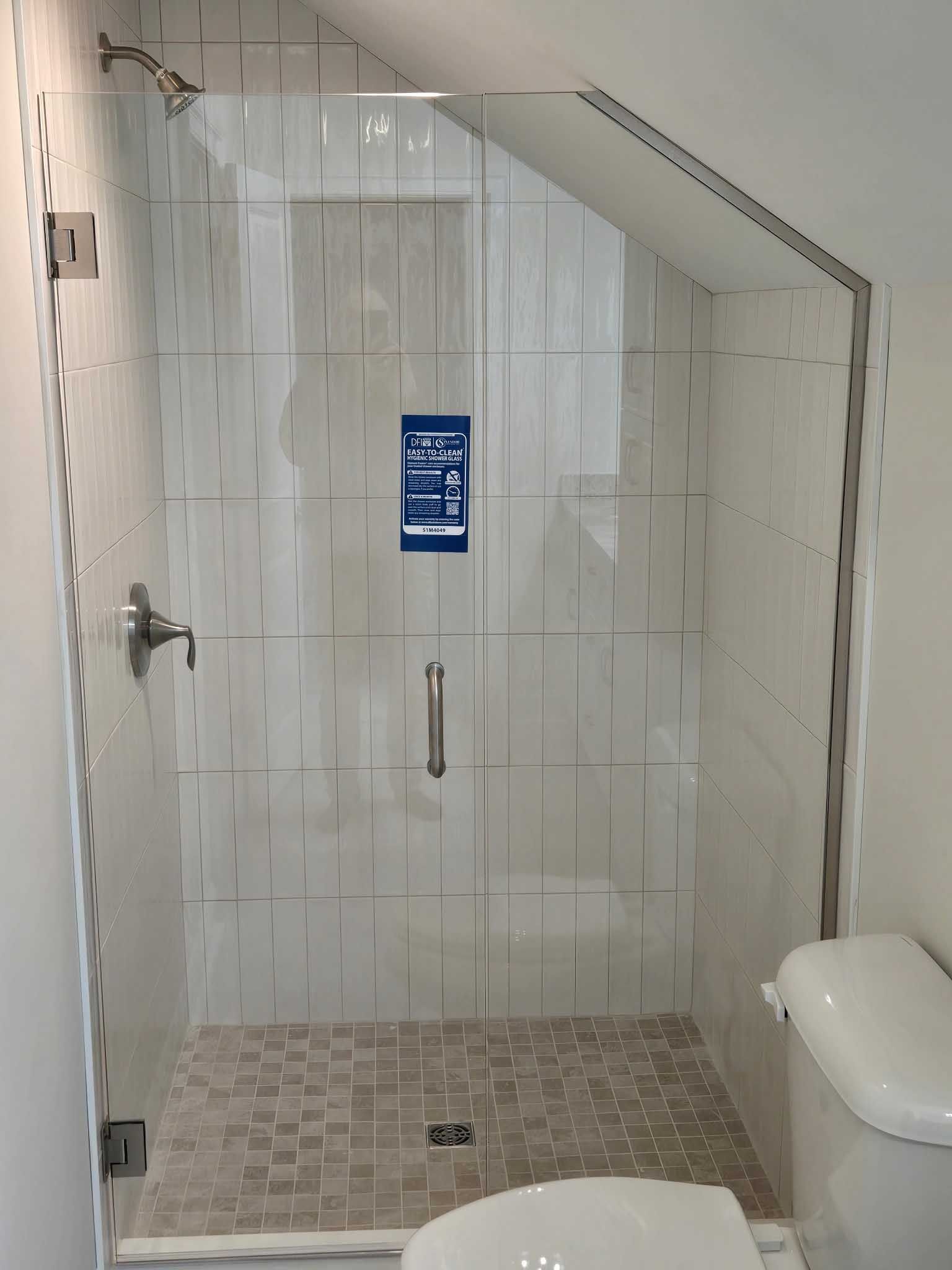 A walk-in shower with a glass door, light-colored tiled walls, and stone mosaic floor, situated next to a white toilet.
