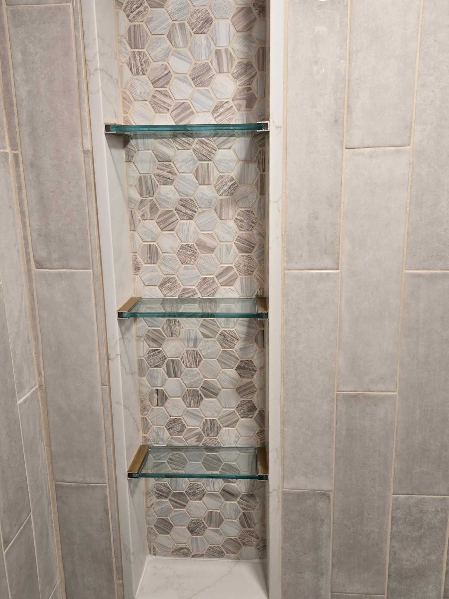 A vertical tiled shower niche with three horizontal glass shelves, set against light gray rectangular wall tiles.