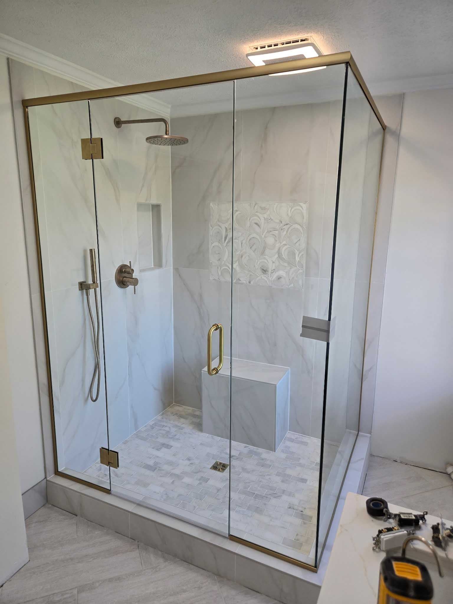 A walk-in shower with gold-toned hardware, white marble-look tiles, a built-in bench, and a glass enclosure in a bathroom.