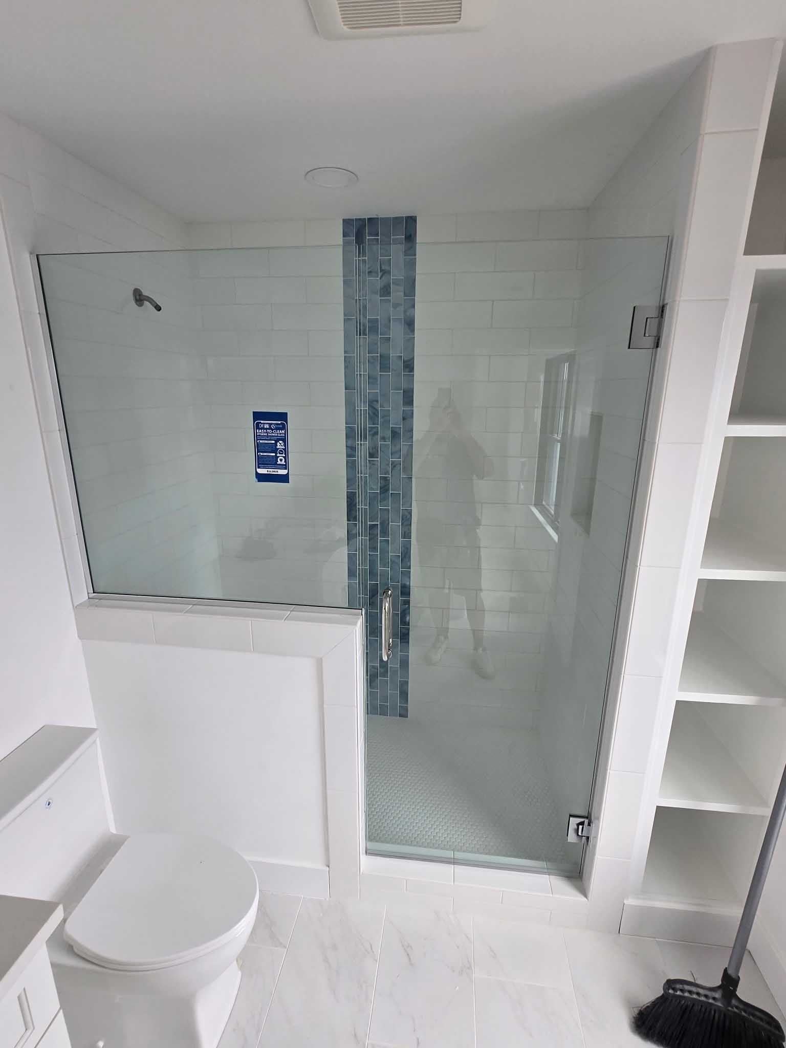 A modern bathroom with a glass shower enclosure featuring a blue tile accent wall, next to a white toilet and open shelves.